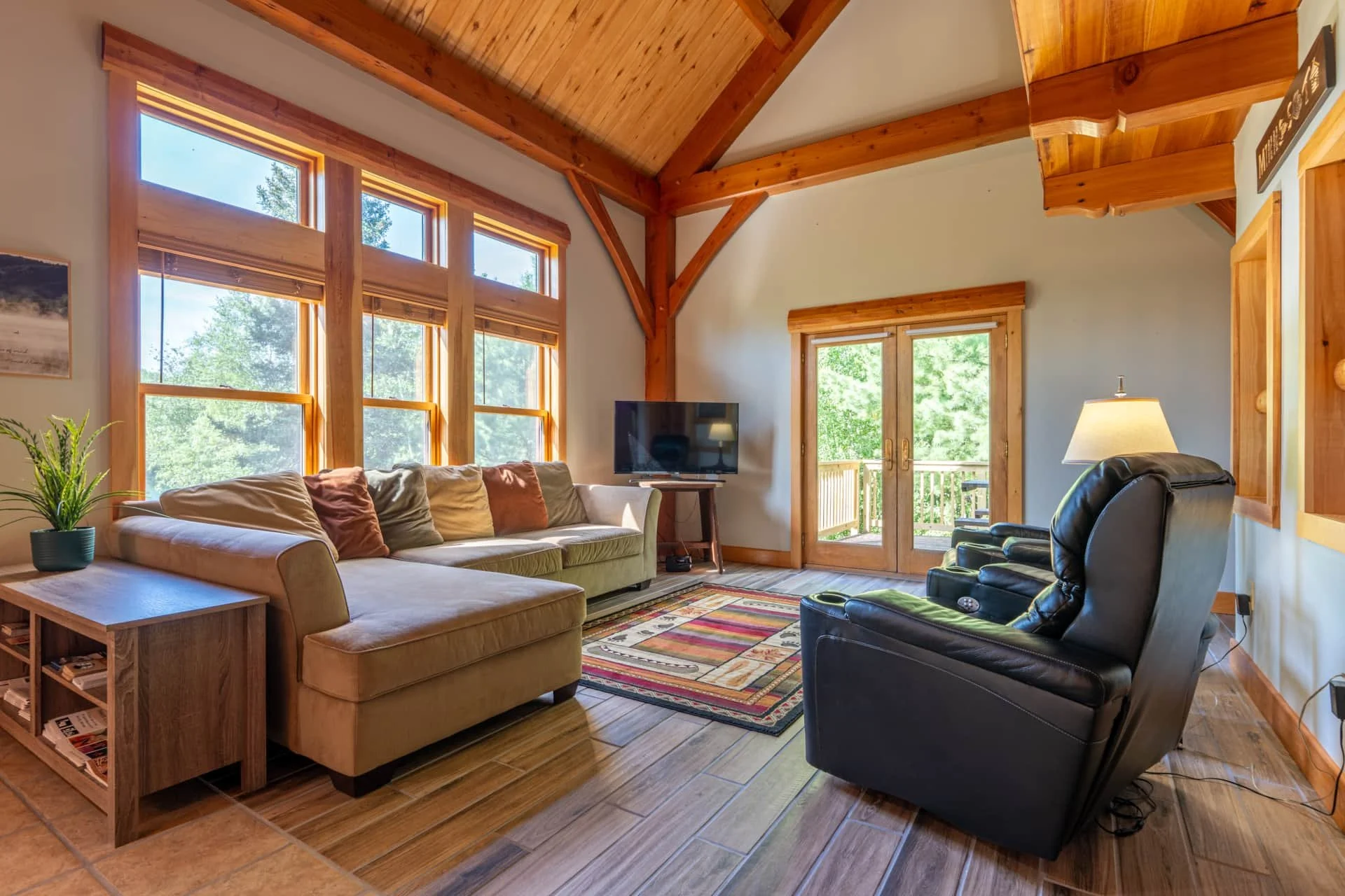 Living room with large windows, wooden ceiling beams, beige sofa with pillows, black leather recliner, wooden side tables, a colorful area rug, potted plant, wall-mounted TV, and sliding glass door leading to a balcony with green trees outside.