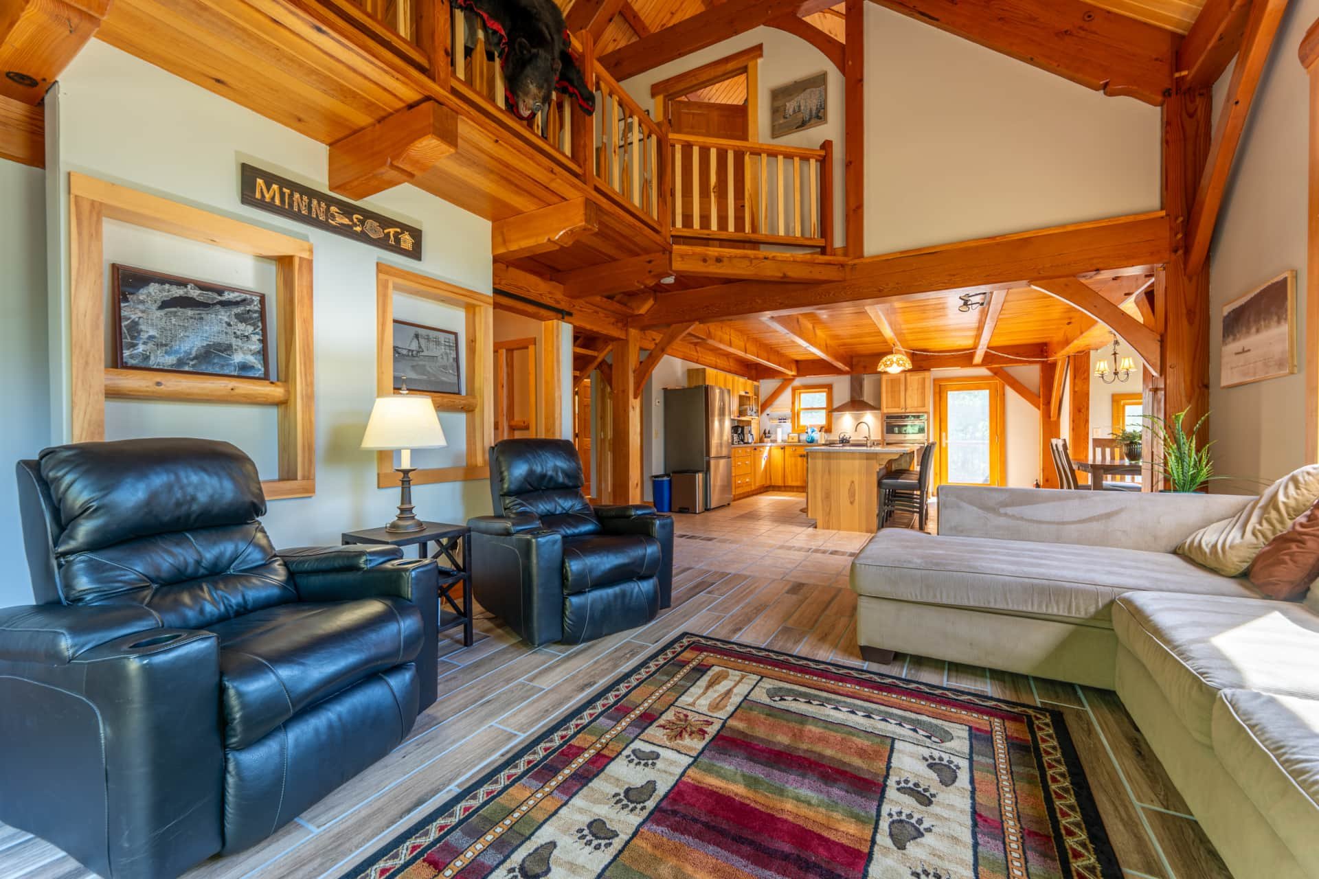 Inside a cozy wooden cabin with a living room, black leather recliner chairs, a beige sofa, a colorful patterned area rug, and a wooden staircase leading to a loft area. The kitchen is visible in the background with stainless steel appliances, wooden