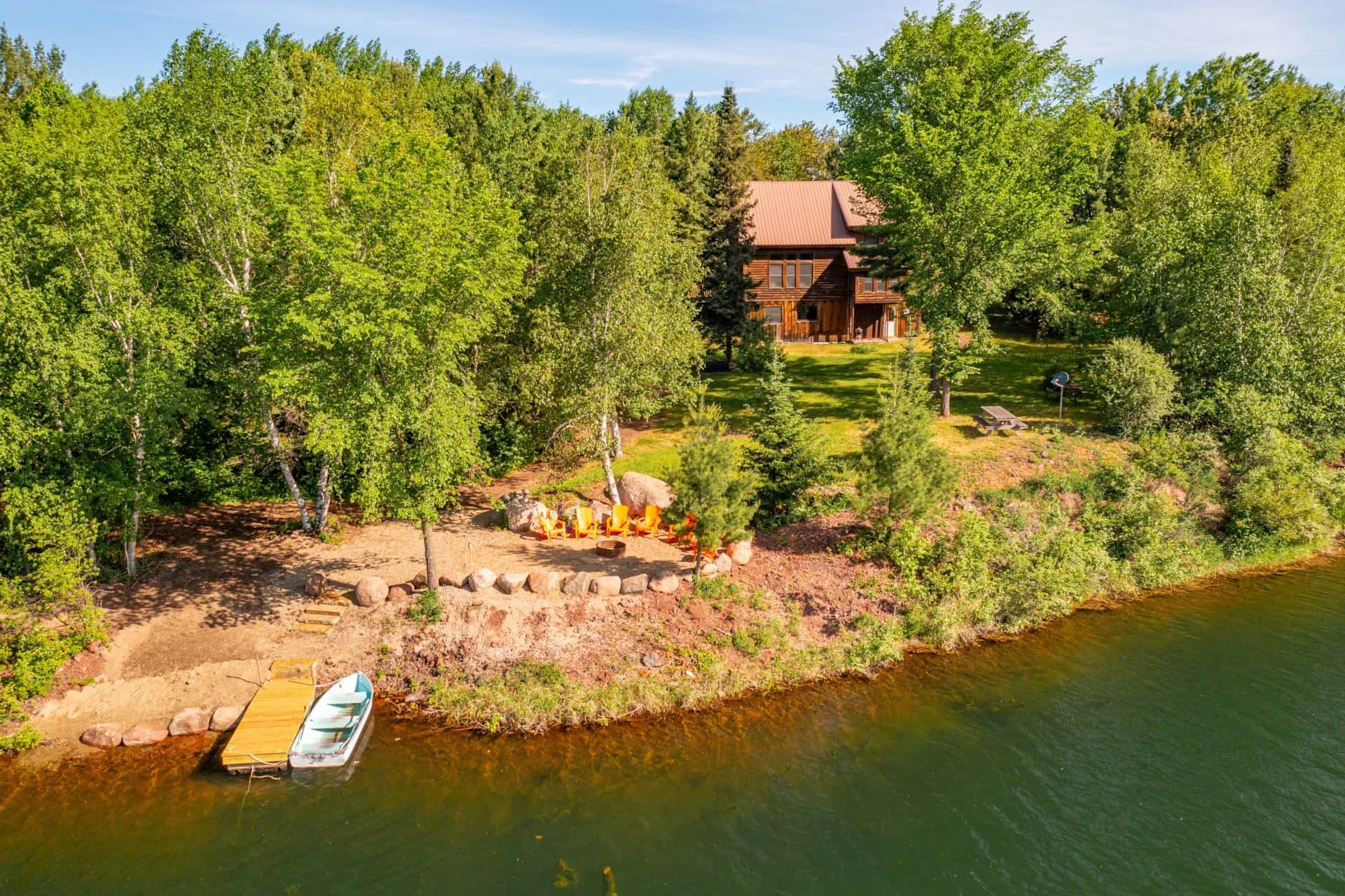 Aerial view of a lakeside house surrounded by green trees, with a sandy beach area featuring orange chairs, a small dock with boats, and a grassy backyard with a picnic table, a satellite dish, and large rocks.