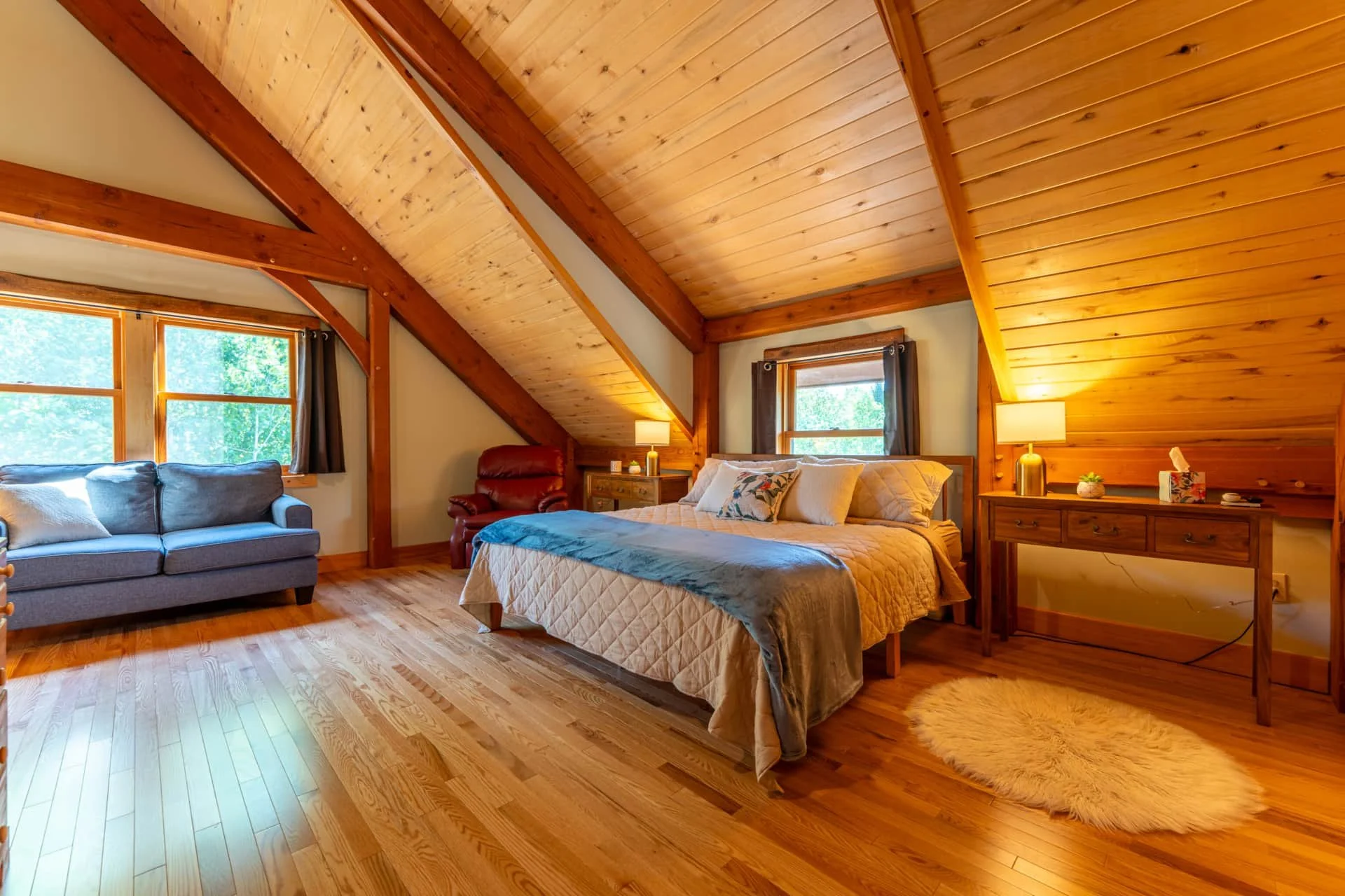 A cozy bedroom with wooden ceilings and floors, featuring a bed, a nightstand with a lamp, a dresser with a plant and tissue box, a window with curtains, a gray sofa, and a red armchair.