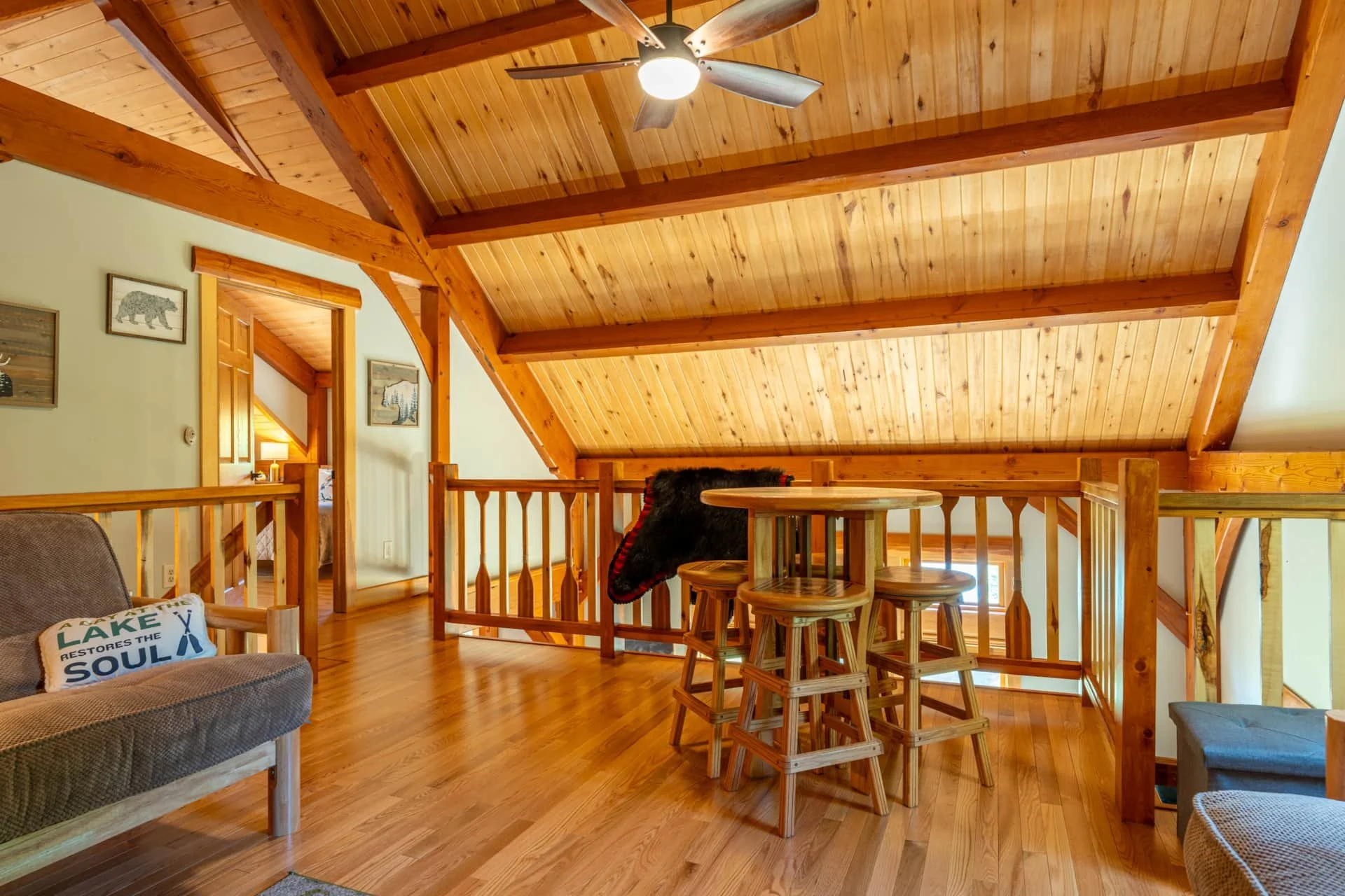 Interior of a cozy cabin with wooden walls and ceiling, featuring a small table with four stools, a couch with a decorative pillow, and framed animal art on the walls.