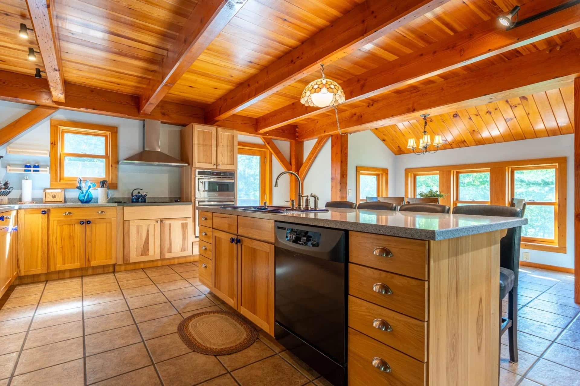 Kitchen with wooden cabinets, a large kitchen island with a sink, stainless steel appliances, and wooden ceiling beams. Multiple windows reveal greenery outside.