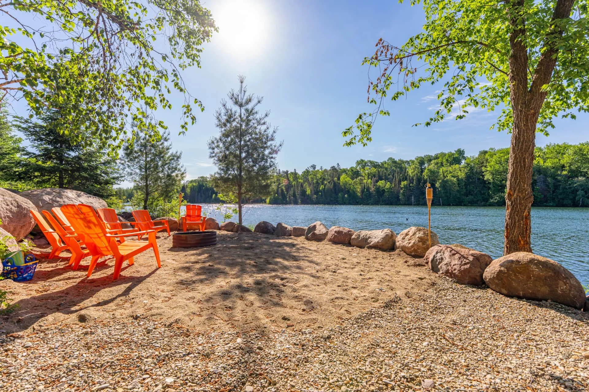 A lakeside scene with orange chairs arranged on a sandy beach, surrounded by large rocks, with green trees and a clear blue sky in the background.