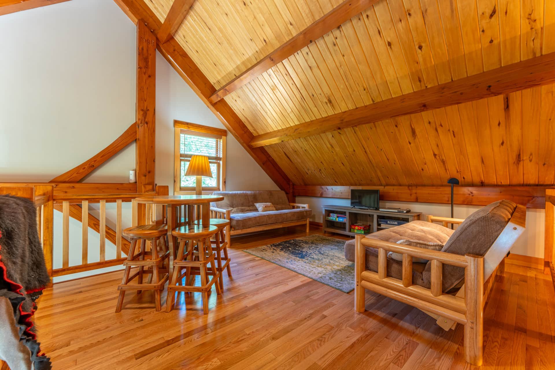 Living room with wooden vaulted ceiling, a window with a lamp, a brown sofa, a flat-screen TV, a bookshelf, and a wooden dining table and stools.