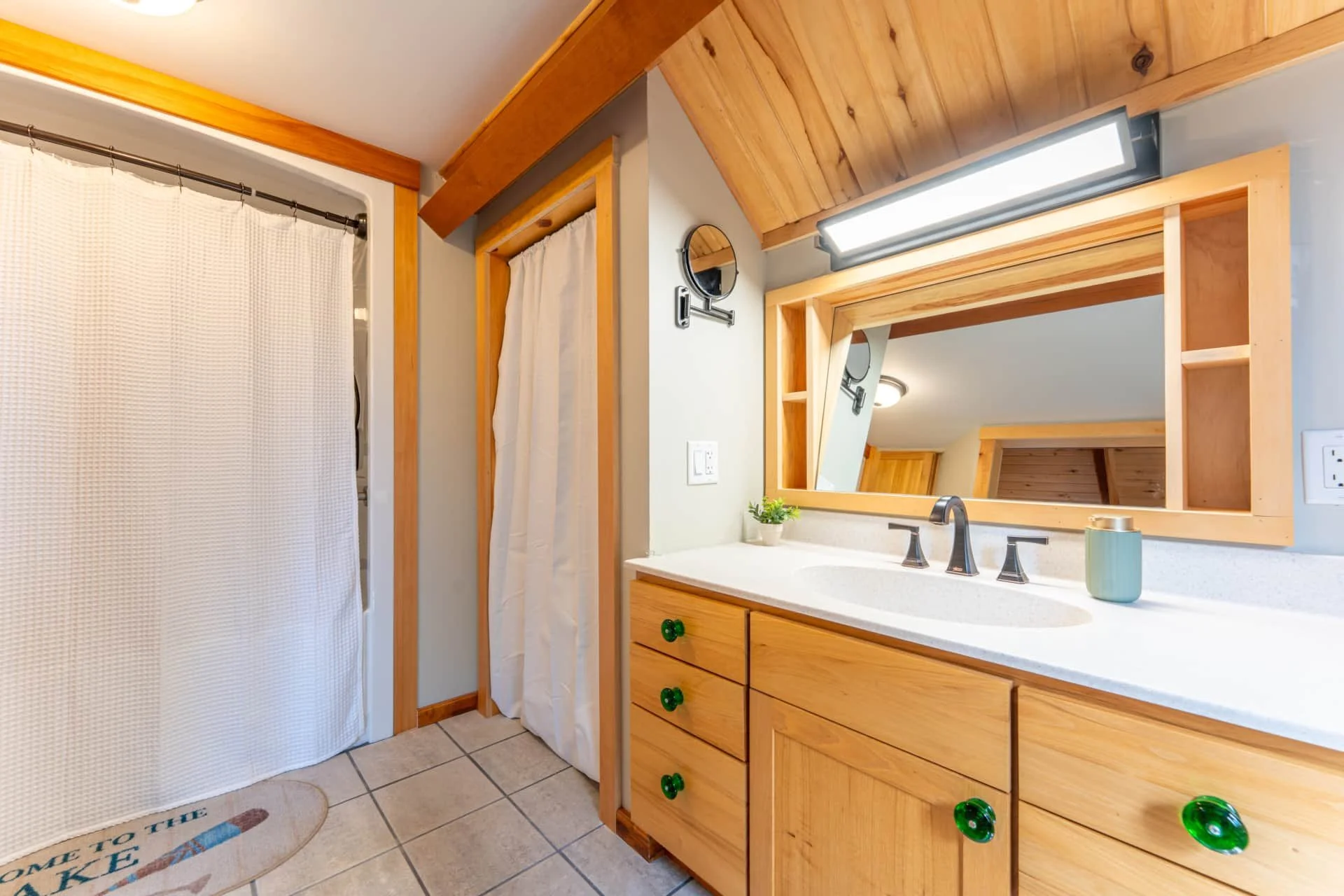 Bathroom vanity with wooden cabinetry and a large mirror, a sink, a small plant, and a soap dispenser. There is a light fixture above the mirror, a small round mirror attached to the wall, and a bathroom door with a white curtain. The flooring is til
