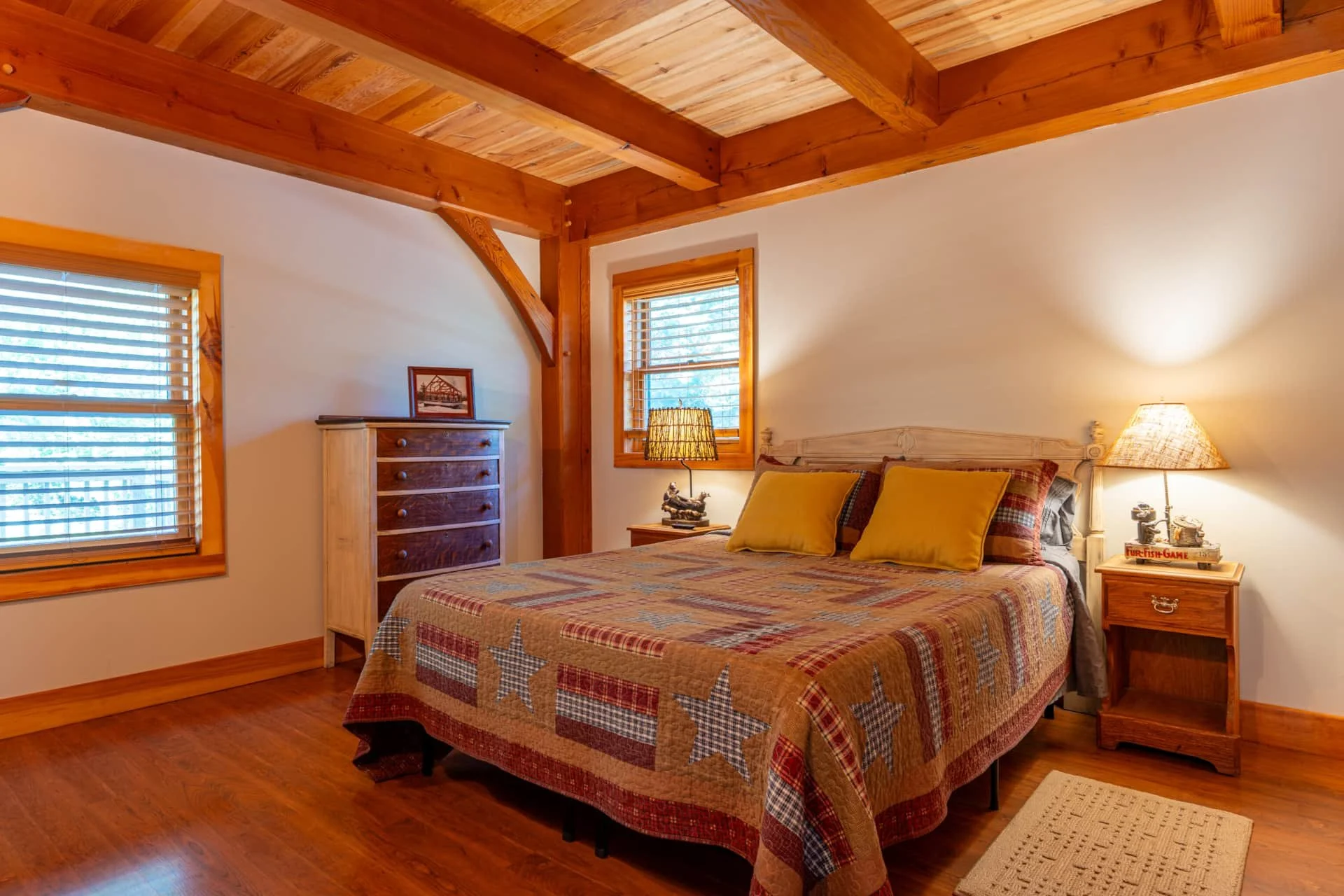 A cozy bedroom with wooden beams and trim, two windows with blinds, a bed with a patchwork quilt, yellow and plaid pillows, a wooden nightstand with a lamp, and a dresser with a framed picture on top.
