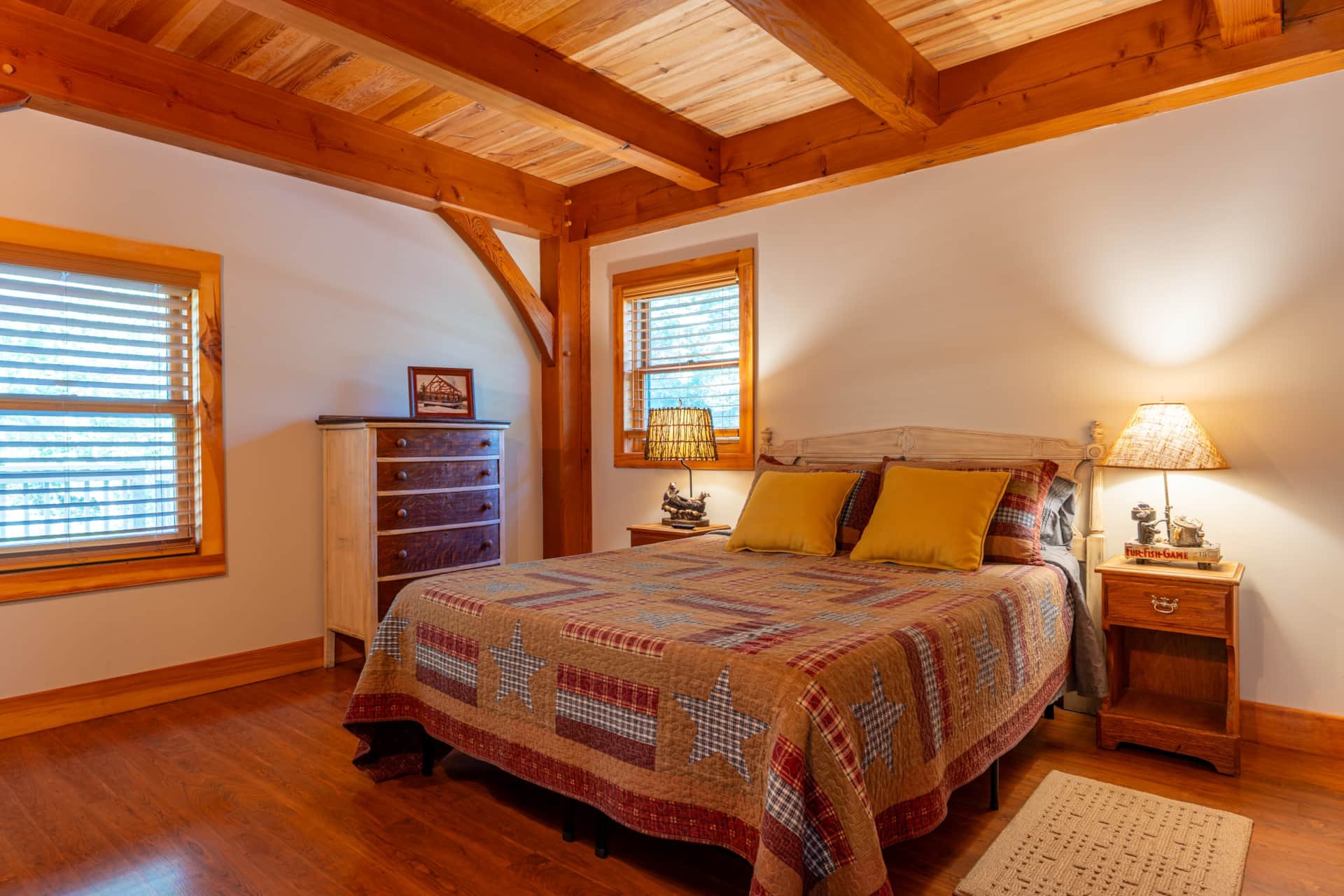 A cozy bedroom with a wooden ceiling and beams, a bed with a quilted bedspread, and two windows with wooden blinds. There are nightstands with lamps and a dresser with a framed photo.