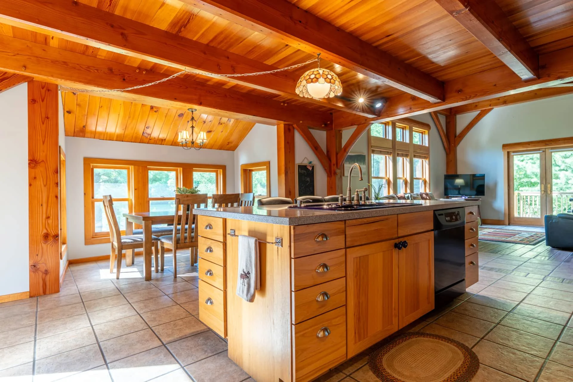  Interior view of a wooden kitchen and dining area with large windows, tiled floor, and a kitchen island with a sink and wooden drawers.