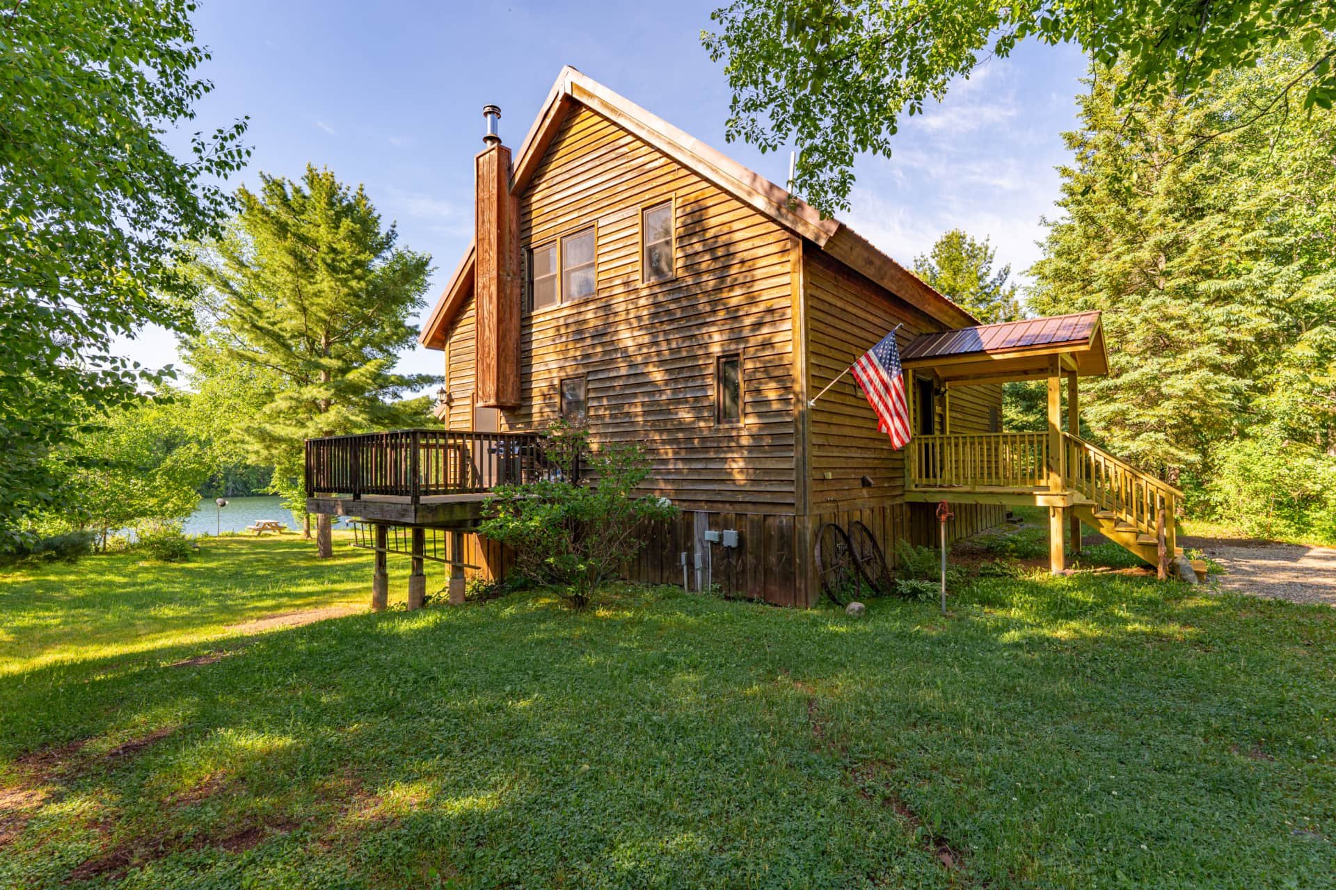Wooden house with an American flag hanging outside, surrounded by trees and grass, near a body of water, under a partly cloudy sky.