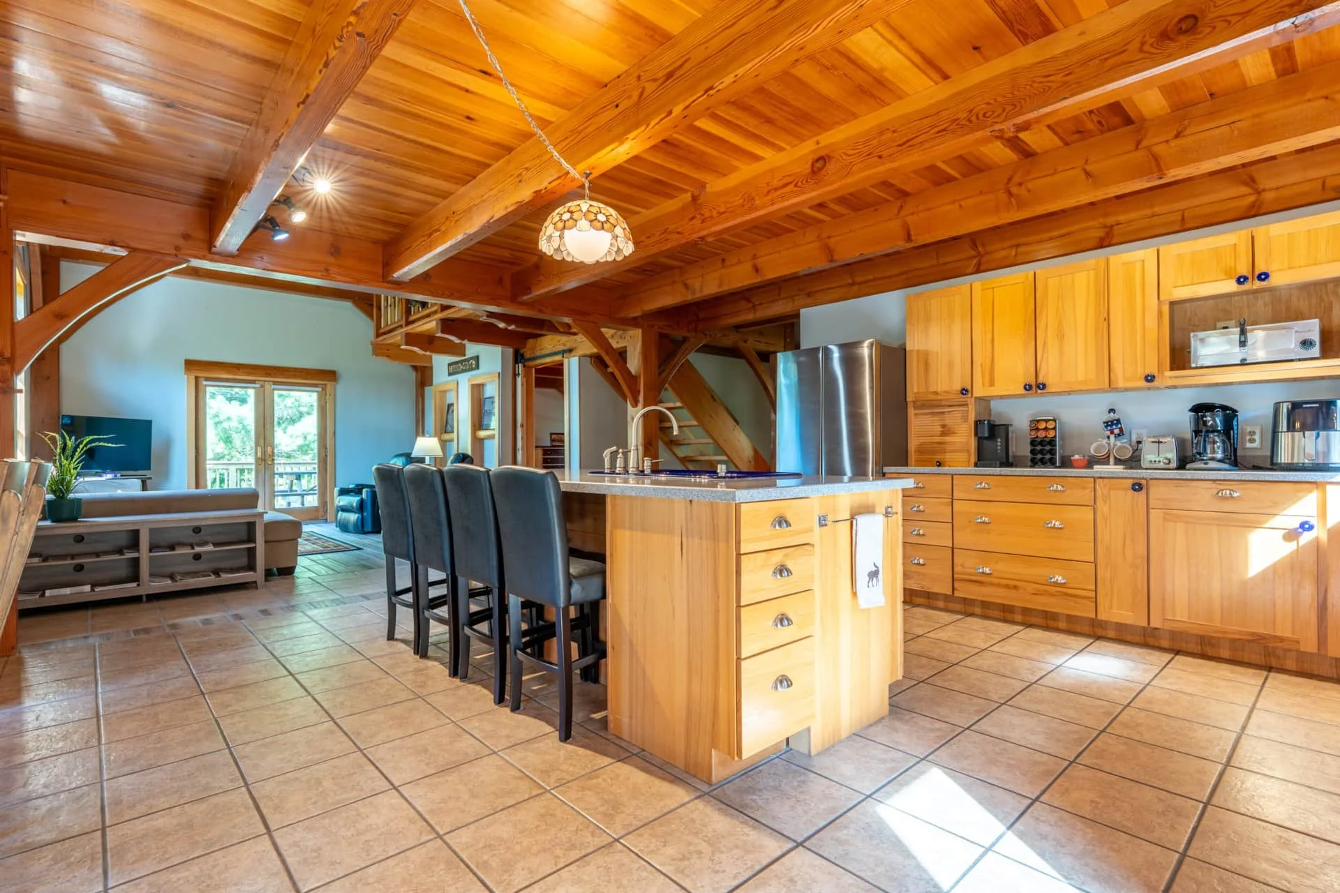 Open-concept kitchen and living room with wooden beams and cabinets, tile flooring, granite countertops, bar stools, stainless steel appliances, and large windows/doors leading outside.