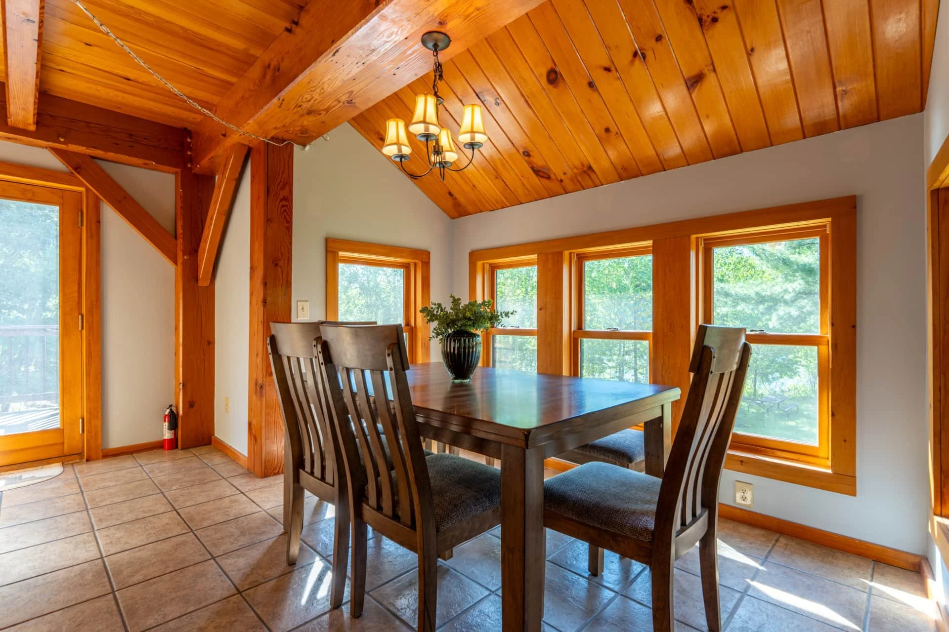 A dining room with a wooden table, six chairs, large windows showing green trees outside, wooden ceiling and window frames, a chandelier with five lamps, a potted plant on the table, and tiled floor.
