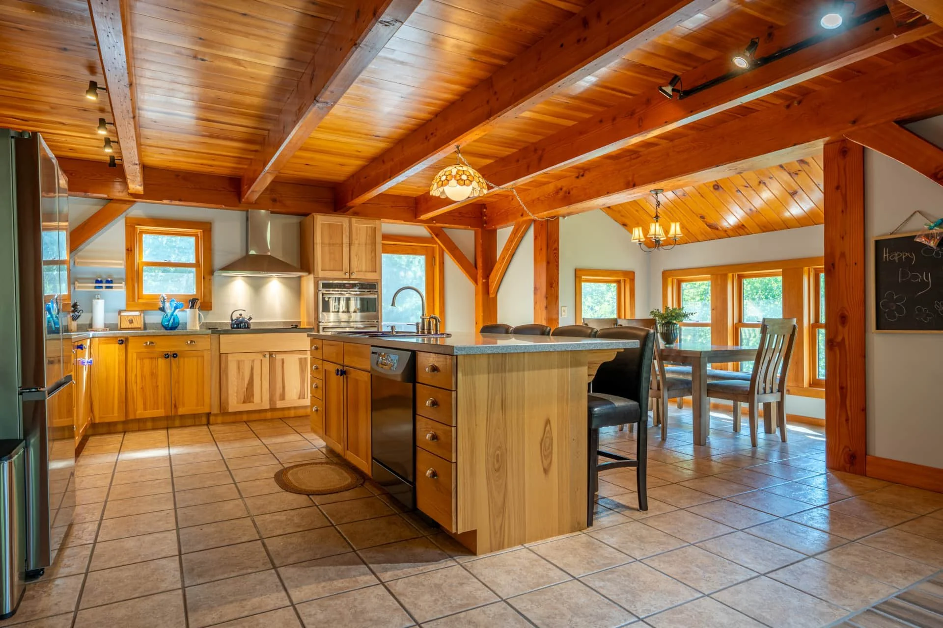 Wooden kitchen with a center island, dining table, and large windows, illuminated by ceiling lights and a chandelier.