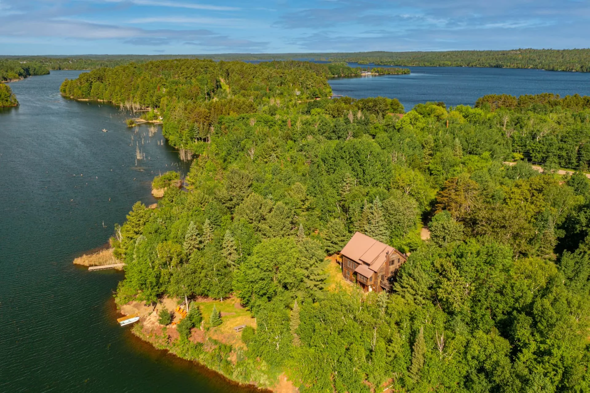 Aerial view of a large lake surrounded by dense green forest with a house near the shoreline and a boat docked in the water.
