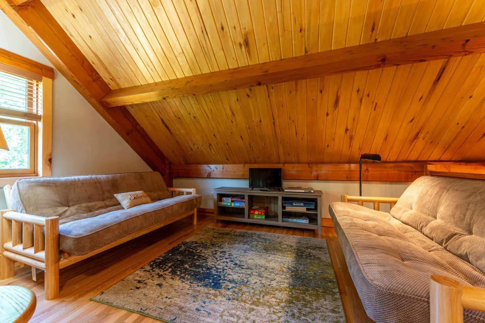 Living room with wooden ceiling and beams, two beige sofas, window with blinds, TV on TV stand, and a patterned area rug.