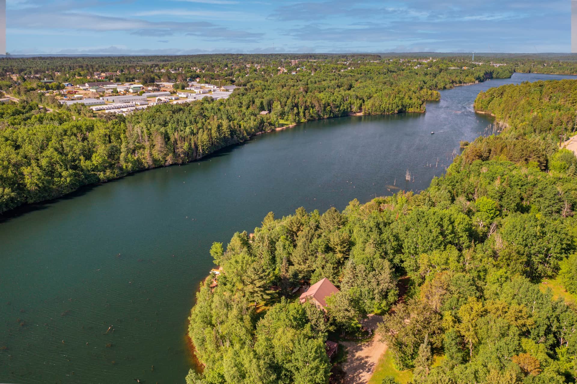 Aerial view of a river flowing through a lush green forest with trees and a few buildings along the shoreline, under a partly cloudy sky.