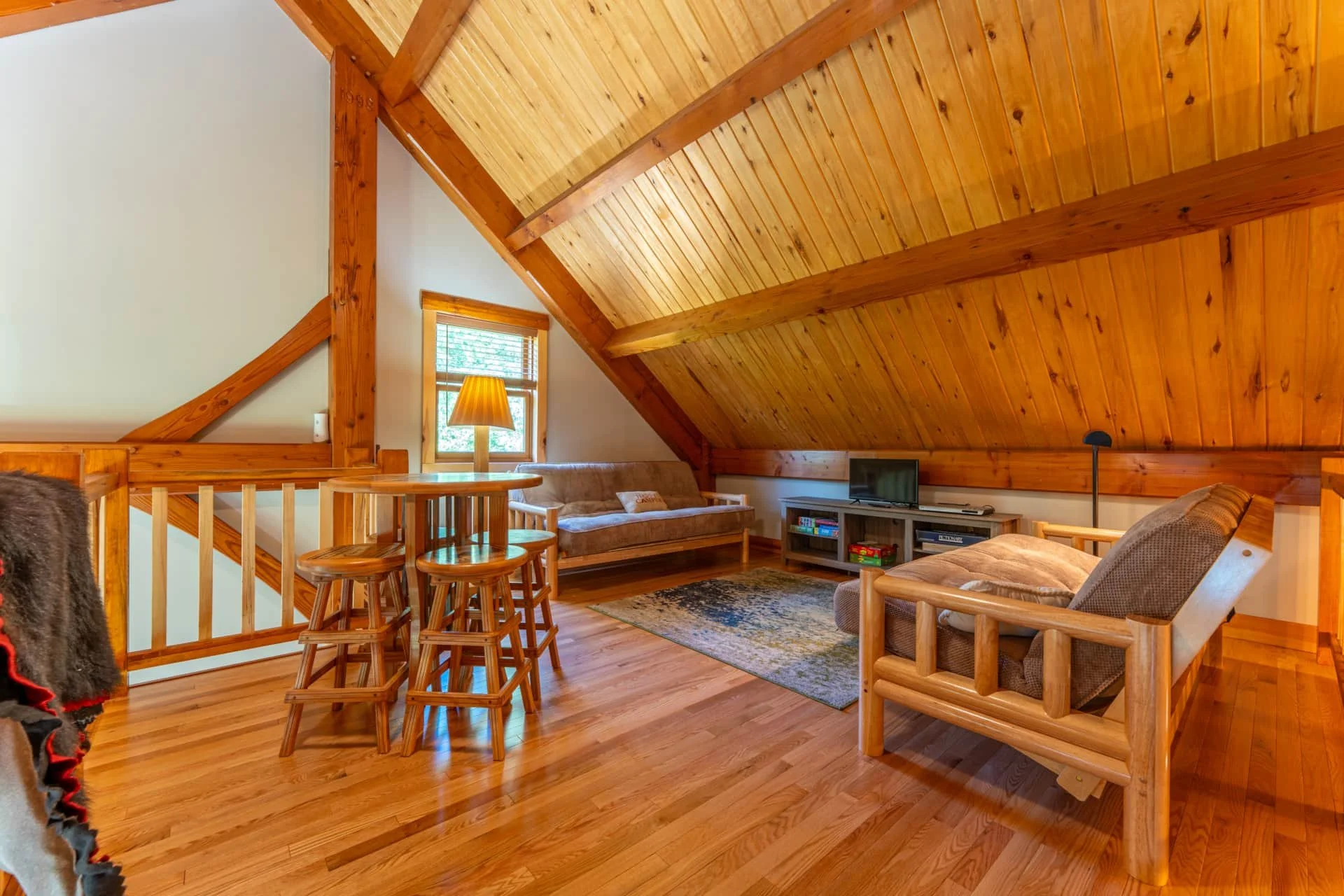 Living room with wooden ceiling and walls, featuring a couch, armchair, small television on a stand, wooden ladder, bar-height wooden table and stools, and window with yellow curtains.