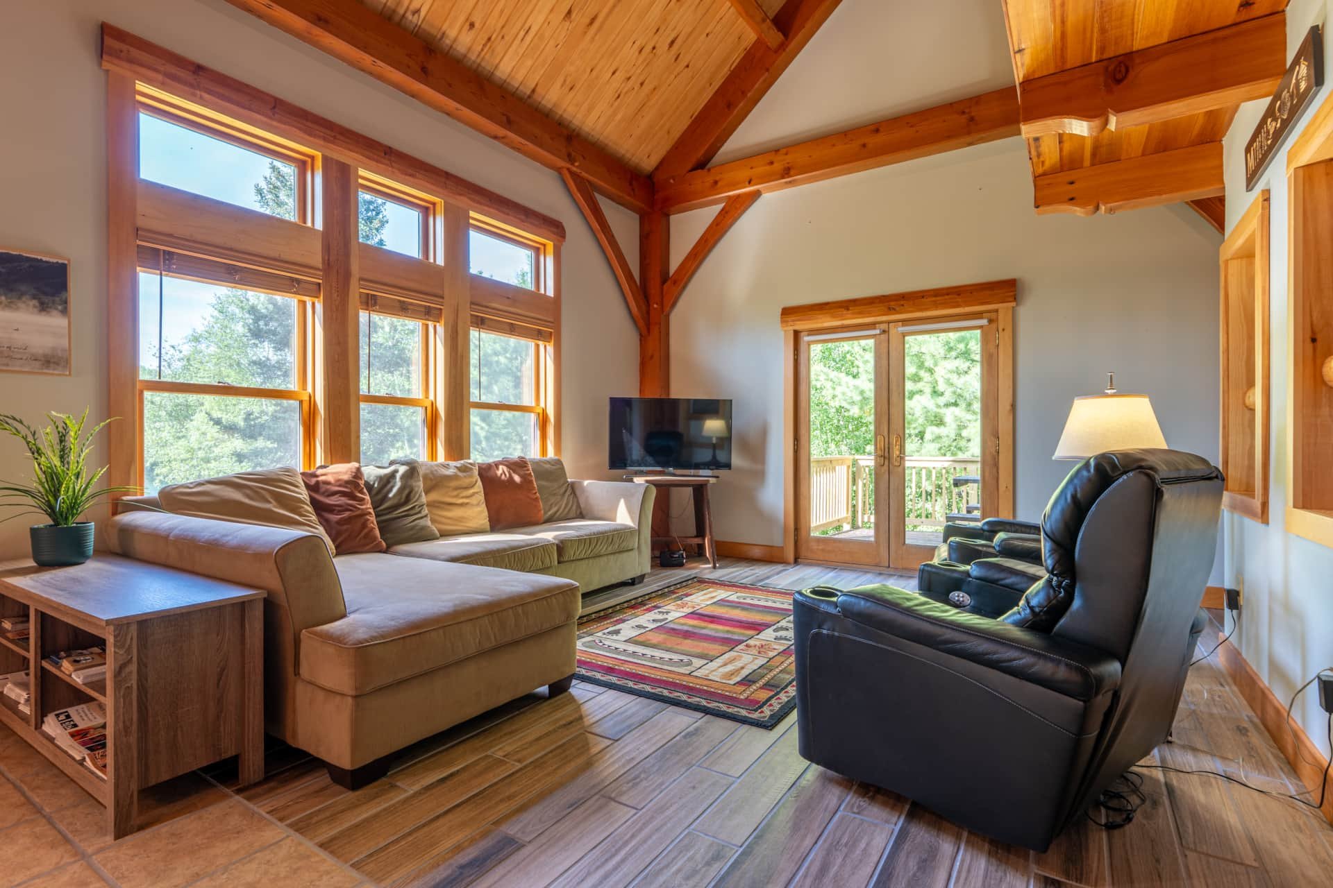 Living room with large windows and sliding glass doors, wooden ceiling accents, cream sofa with pillows, black recliner, wooden side table, television, colorful area rug, potted plant, and a lamp.