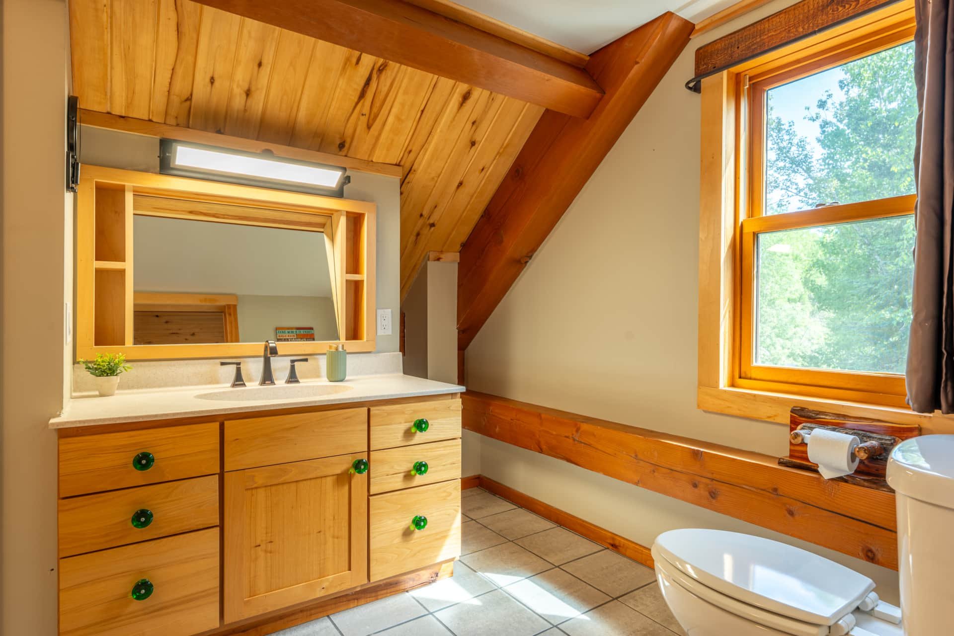 Bathroom with wooden ceiling and window, wooden vanity with mirror, small plant and soap dispenser, and a toilet, with sunlight coming through the window.