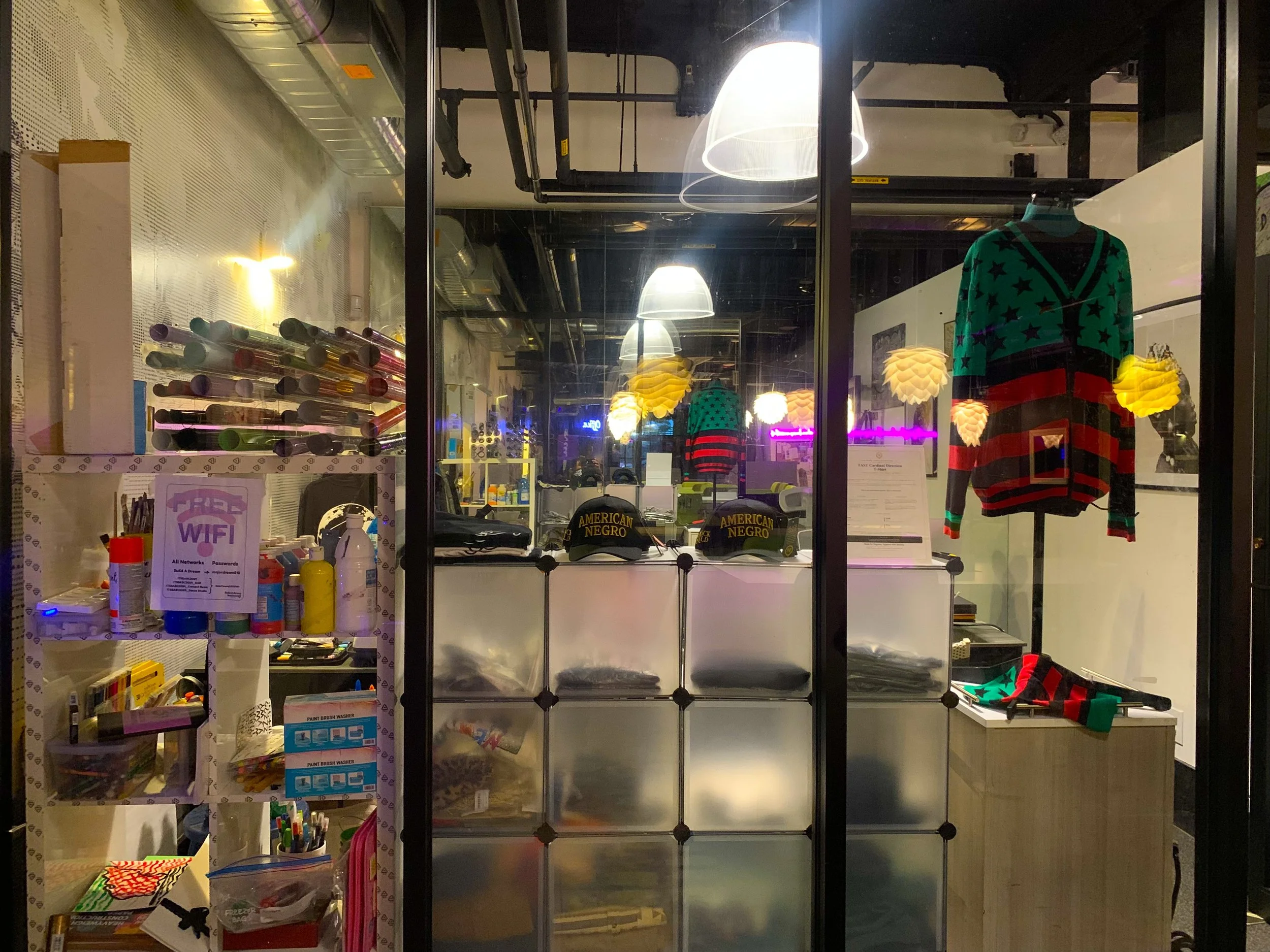 A store window display with various items including clothing, hats, and accessories, seen through the glass with reflections and interior lighting.