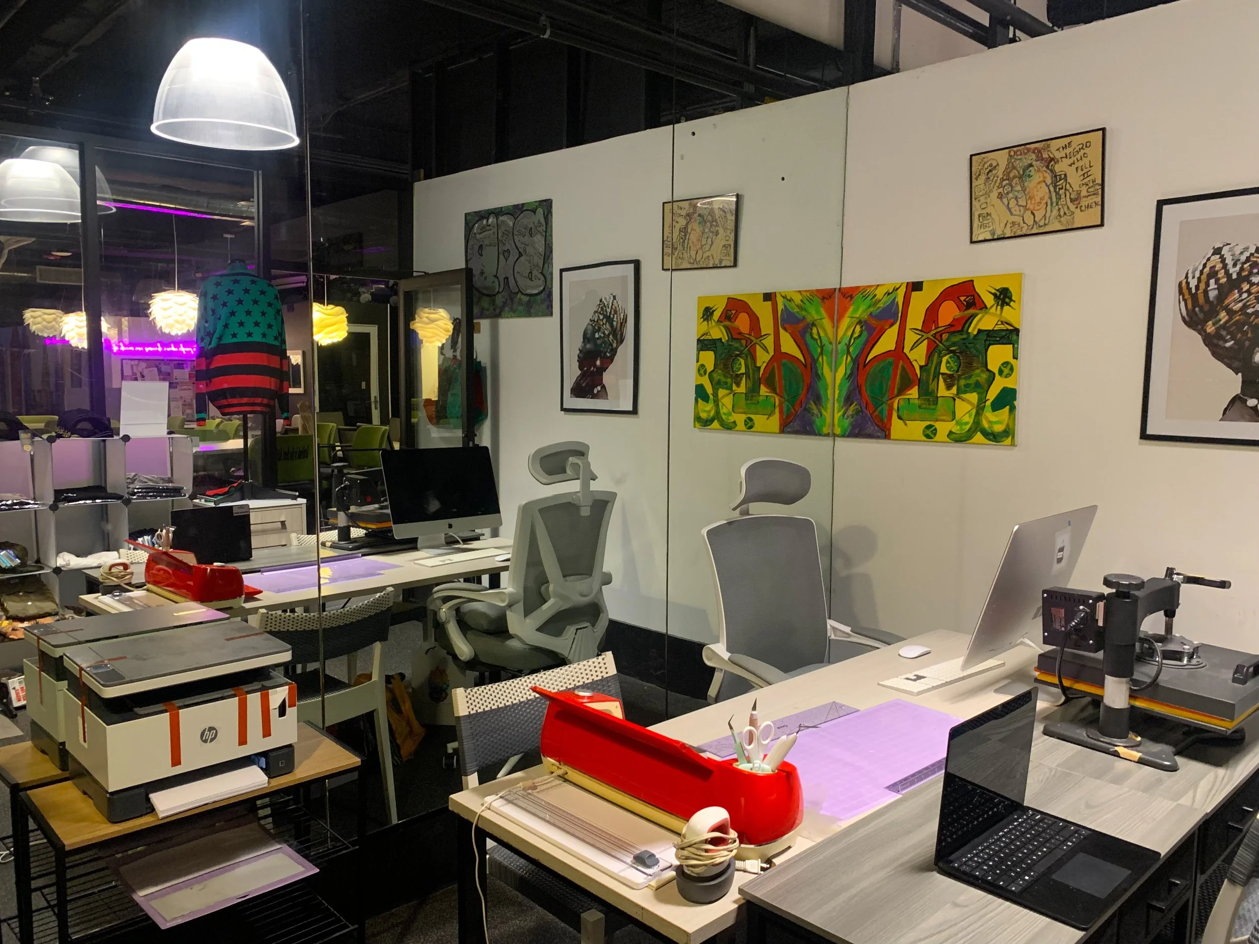 An office or art gallery space with diverse artwork on white walls, multiple desks with computers, office supplies, and chairs, illuminated by large pendant lights, with reflections of pink neon light in the glass window.
