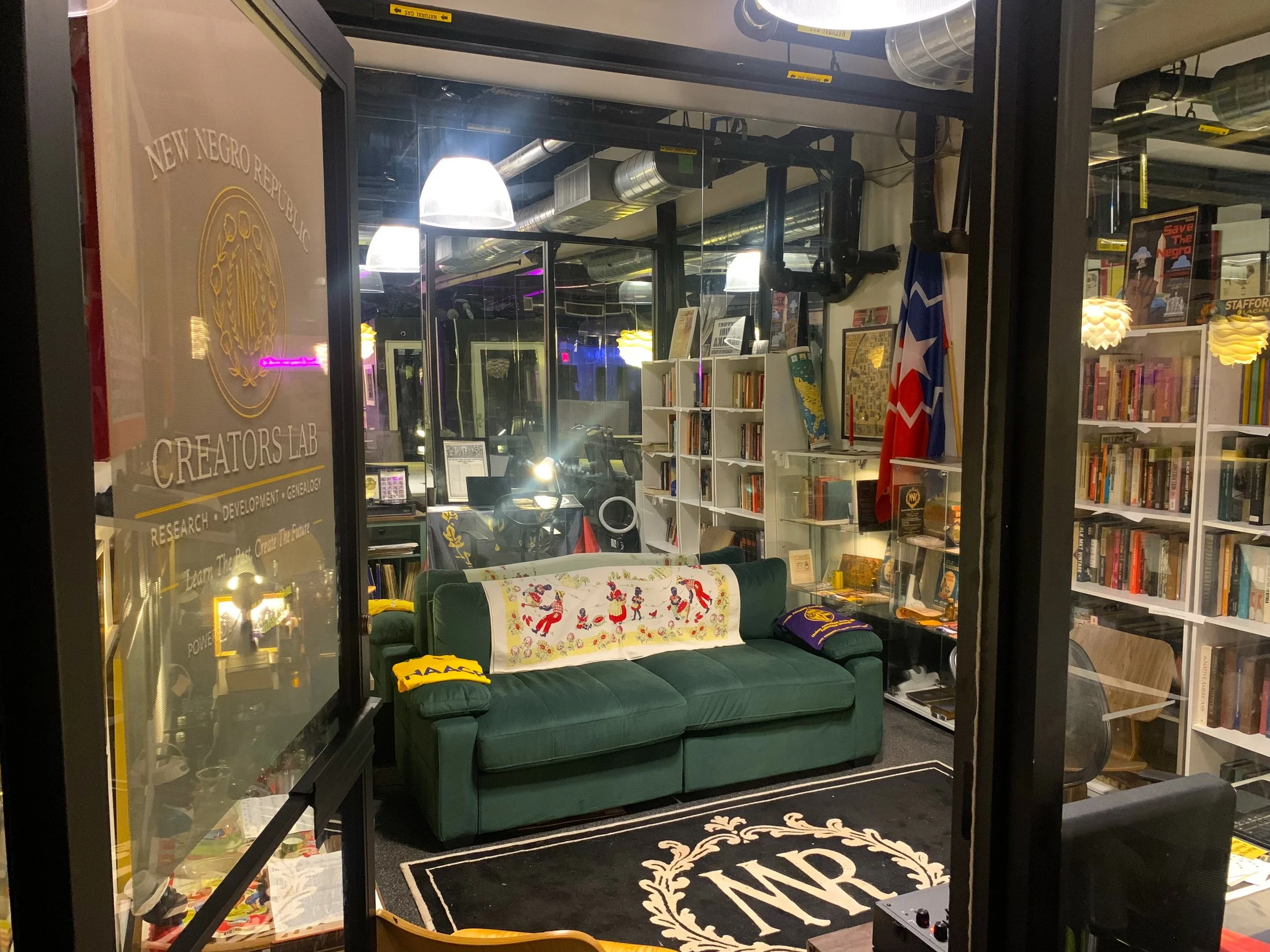 View of a bookstore or library with shelves of books, a green velvet sofa with a decorative cloth, flags, and various lighting fixtures.
