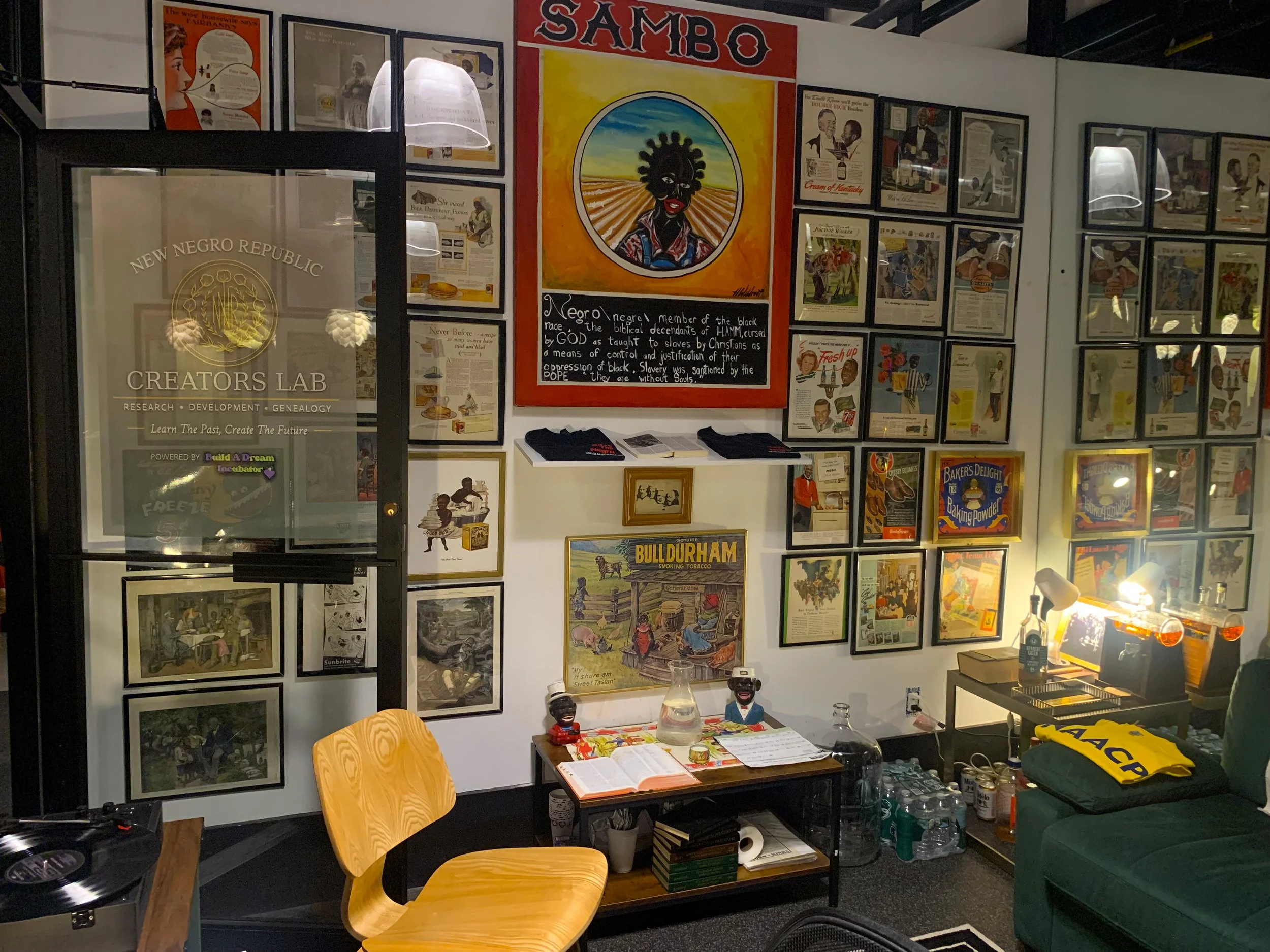 A wall filled with framed posters and artwork, including colorful illustrations and vintage ads, with a prominent red poster featuring a black woman with afro hairstyle and a quote about race and slavery. A small table in front holds books, papers, and decorative figurines, and there's a glass of water on the table.