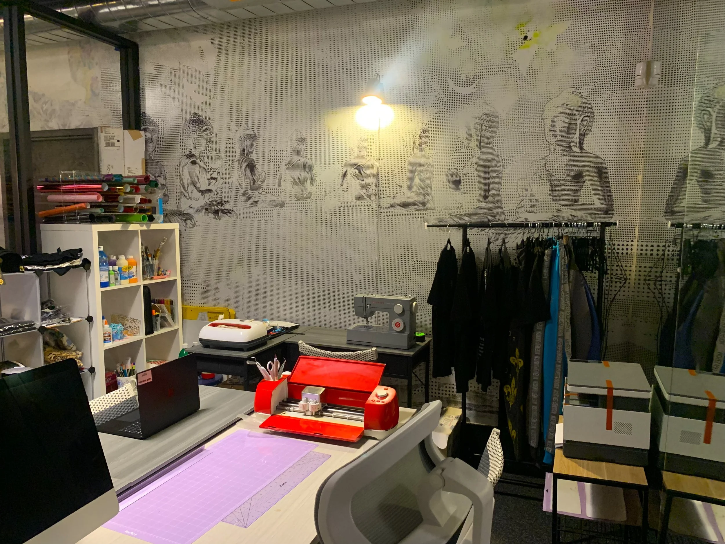 A cluttered workspace with a table, a computer, a red construction tool, and various office and craft supplies. A wall in the background has a mural of people sitting in a line. There are shelves with boxes, tubes, and bottles. Clothing hangs on a rack, and a sewing machine is on the table.