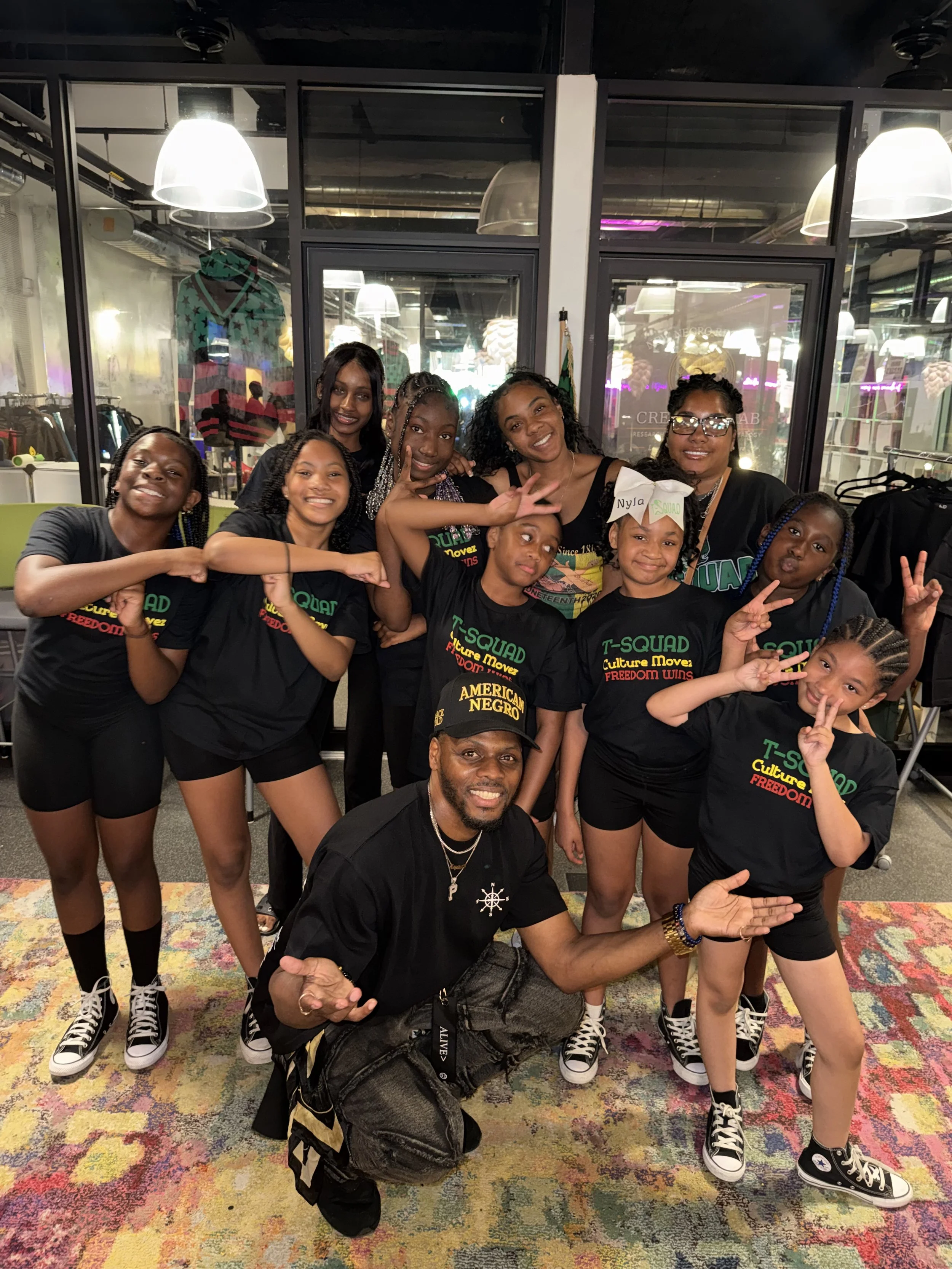 A group of ten people, mostly young girls, posing for a photo indoors. They are smiling and making peace signs or other hand gestures. The group is wearing coordinated black T-shirts with colorful text that says 'T-SQUAD' and other words. Some girls have braided hair, and one girl is wearing glasses. There is a man kneeling in the front wearing a black cap and shirt, with a colorful backpack on the floor. The background features glass walls, clothing racks, and colorful lighting.
