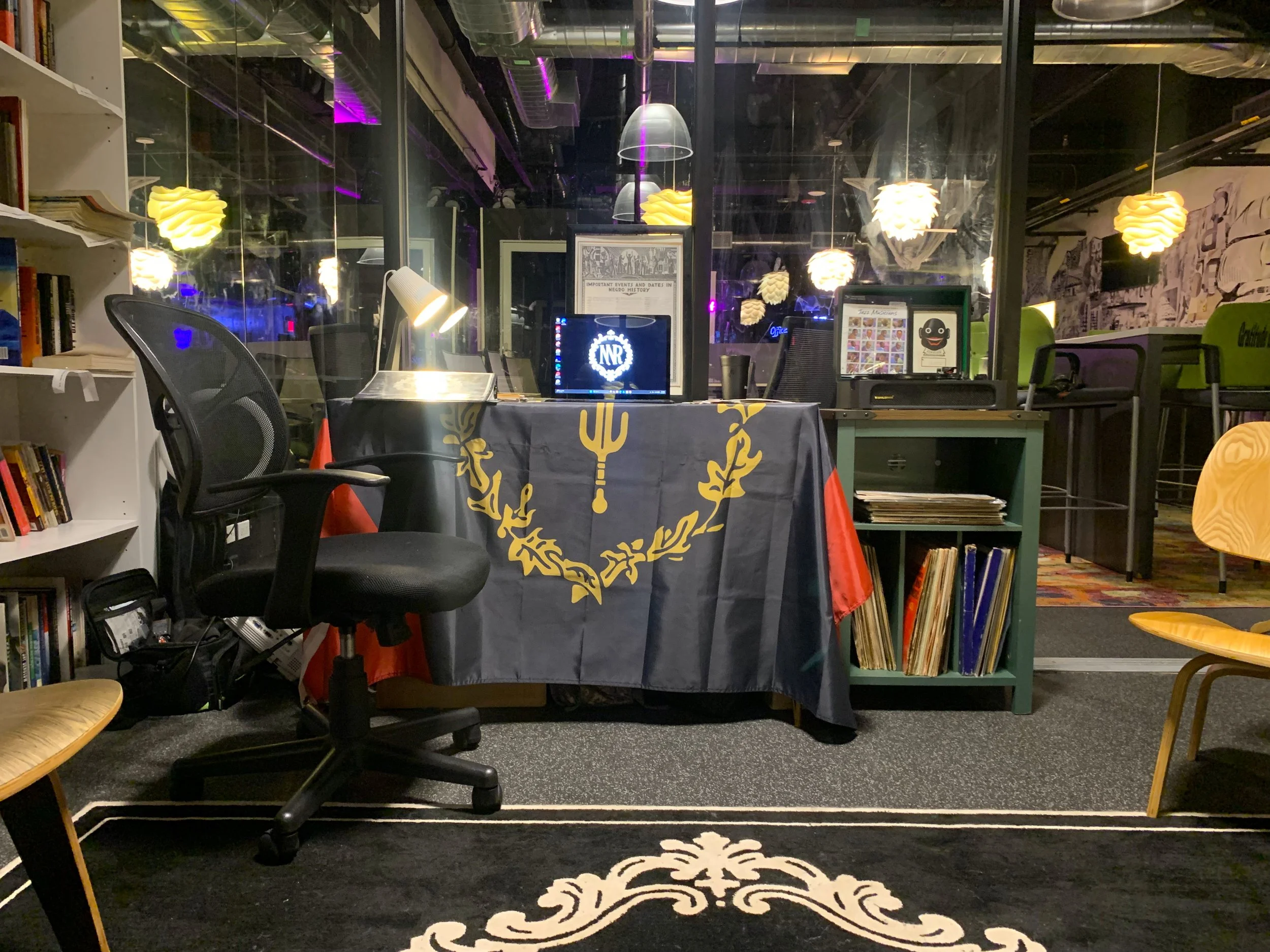 An indoor workspace with a table covered by a black cloth with gold designs, surrounded by black and yellow chairs, and decorated with decorative yellow hanging lights. There are bookshelves on the left and a glass wall with a reflection of the lights. In the center, there is a computer monitor, a desk lamp, and framed pictures. The background includes stacked chairs and a colorful floor and wall mural.
