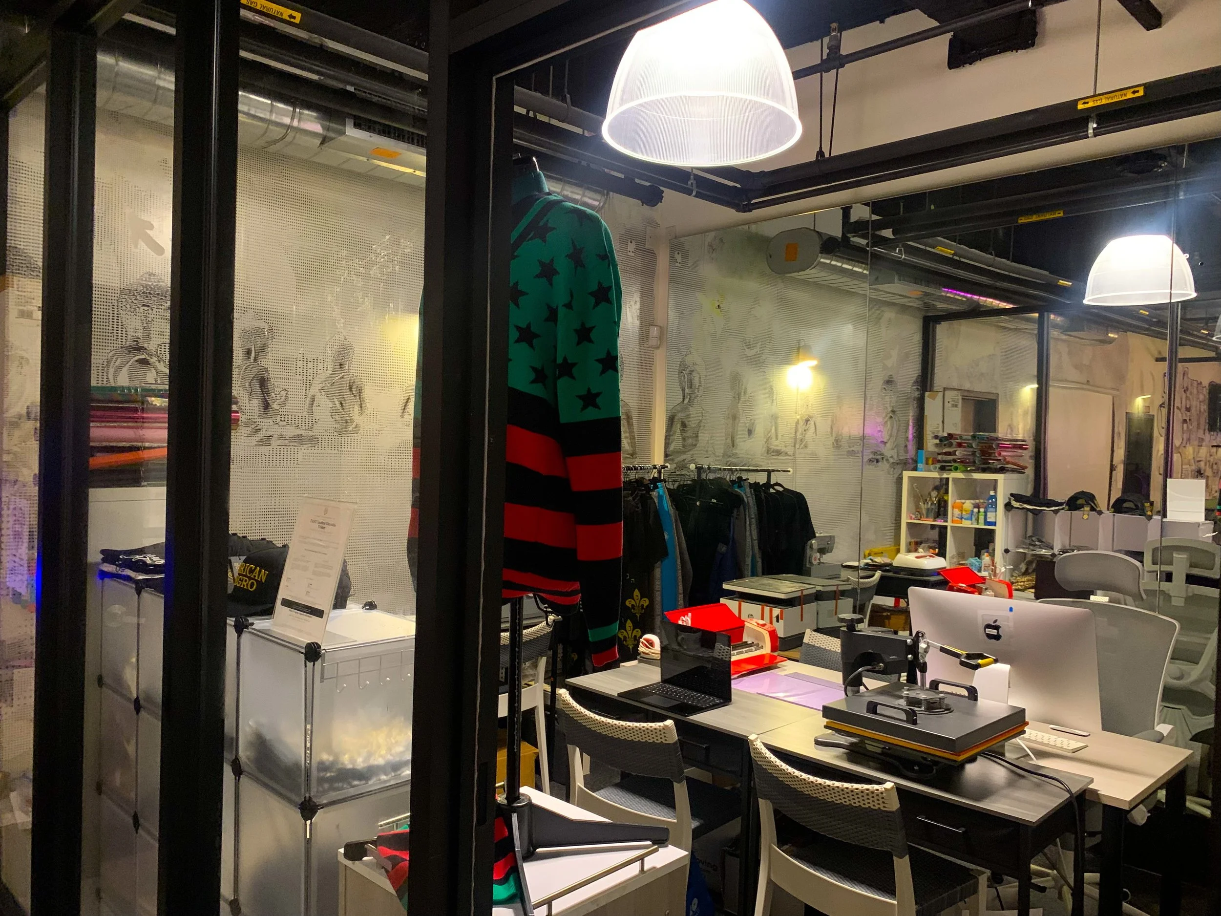 Inside a retail store with glasses partition walls, a mannequin dressed in a rainbow-colored sweater with star patterns, a desk with a laptop, and various shelves with objects and books. Bright overhead lights illuminate the area.