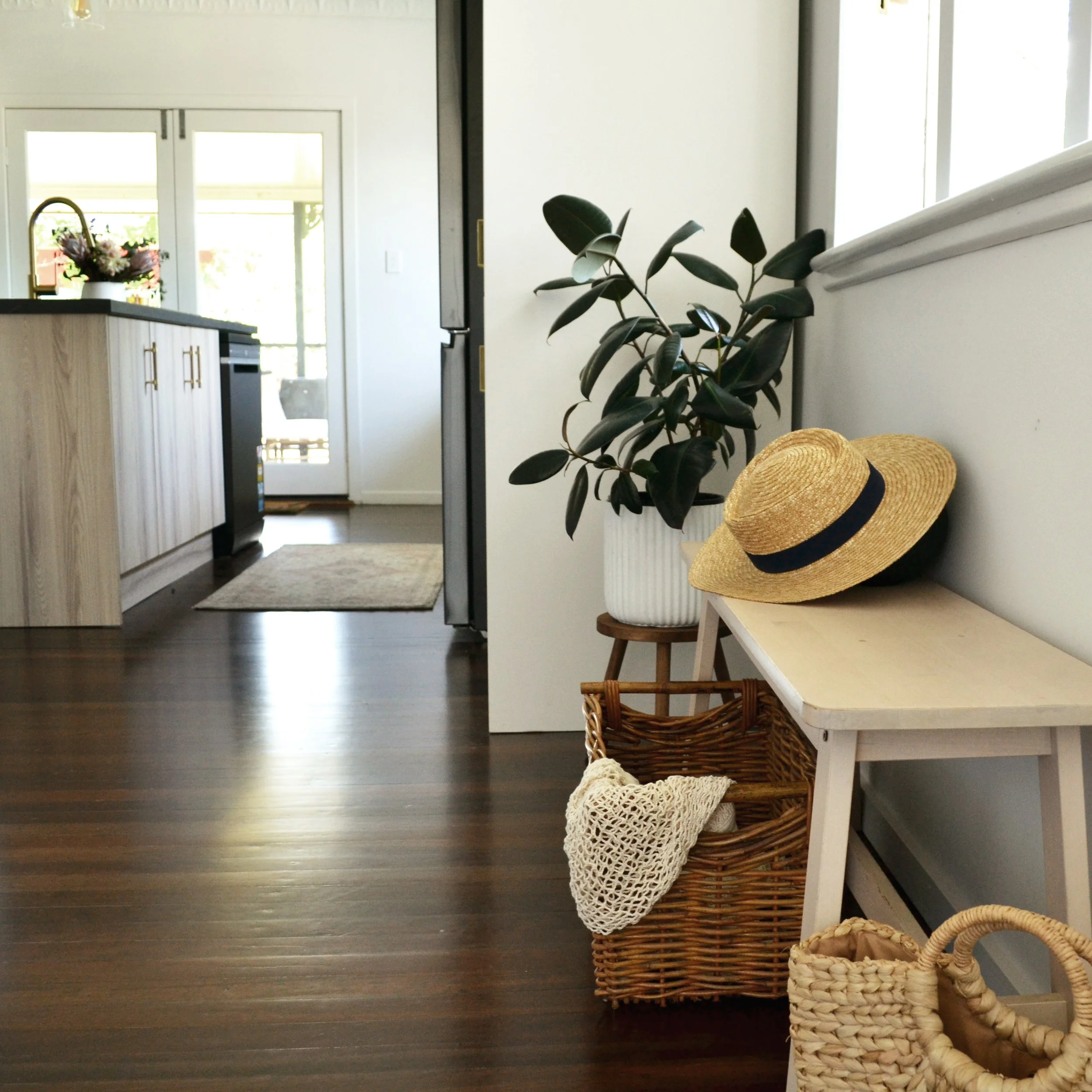 A room with dark wooden flooring, a white dresser with gold handles, a potted plant with dark green leaves, a woven straw hat and a small woven basket on a white piece of furniture, and a window letting in bright natural light.