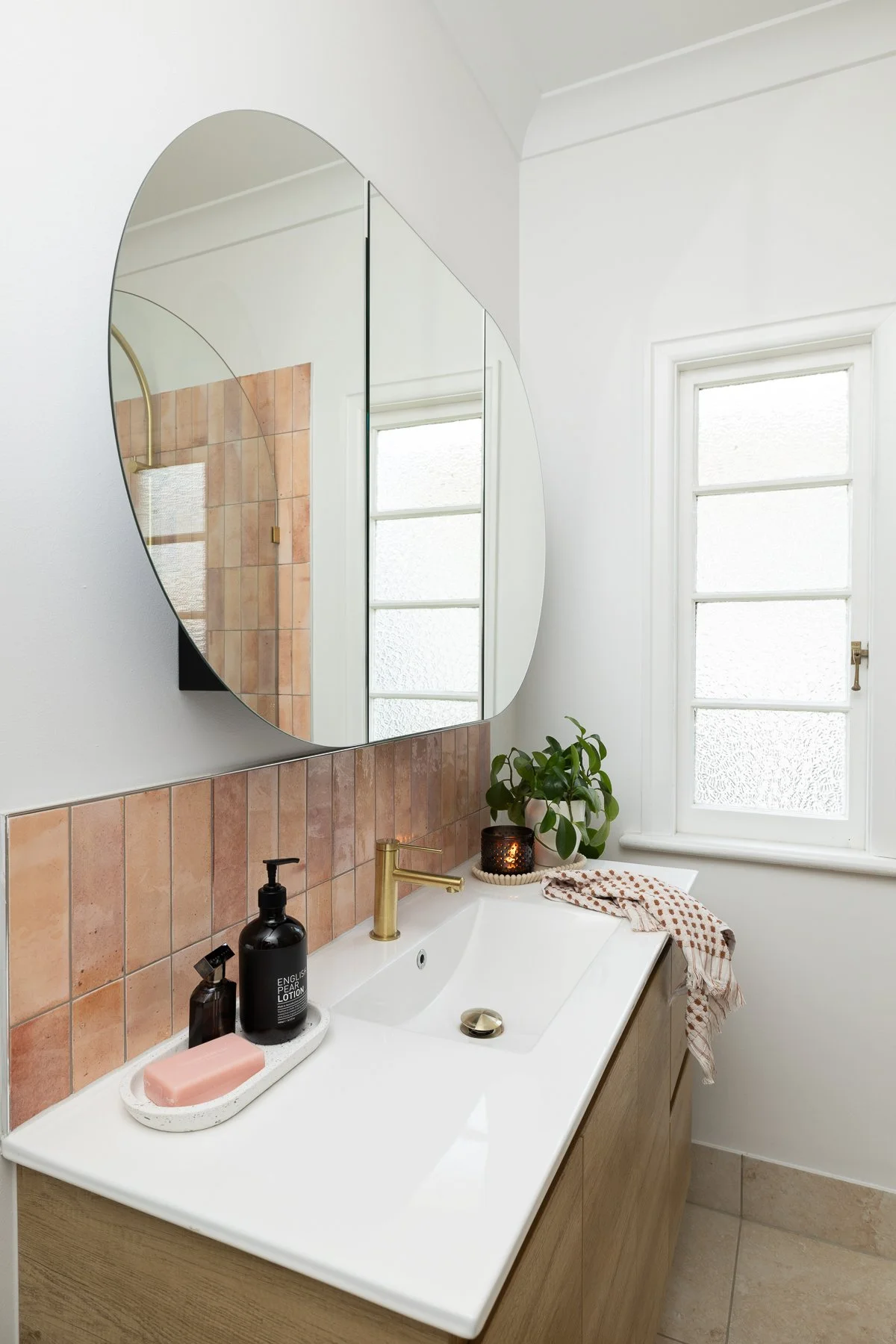 A bathroom sink area with a large, rounded mirror, a window with frosted glass, a brass faucet, a potted plant, a candle, soap, and lotion on a tray, and a towel hanging over the sink.
