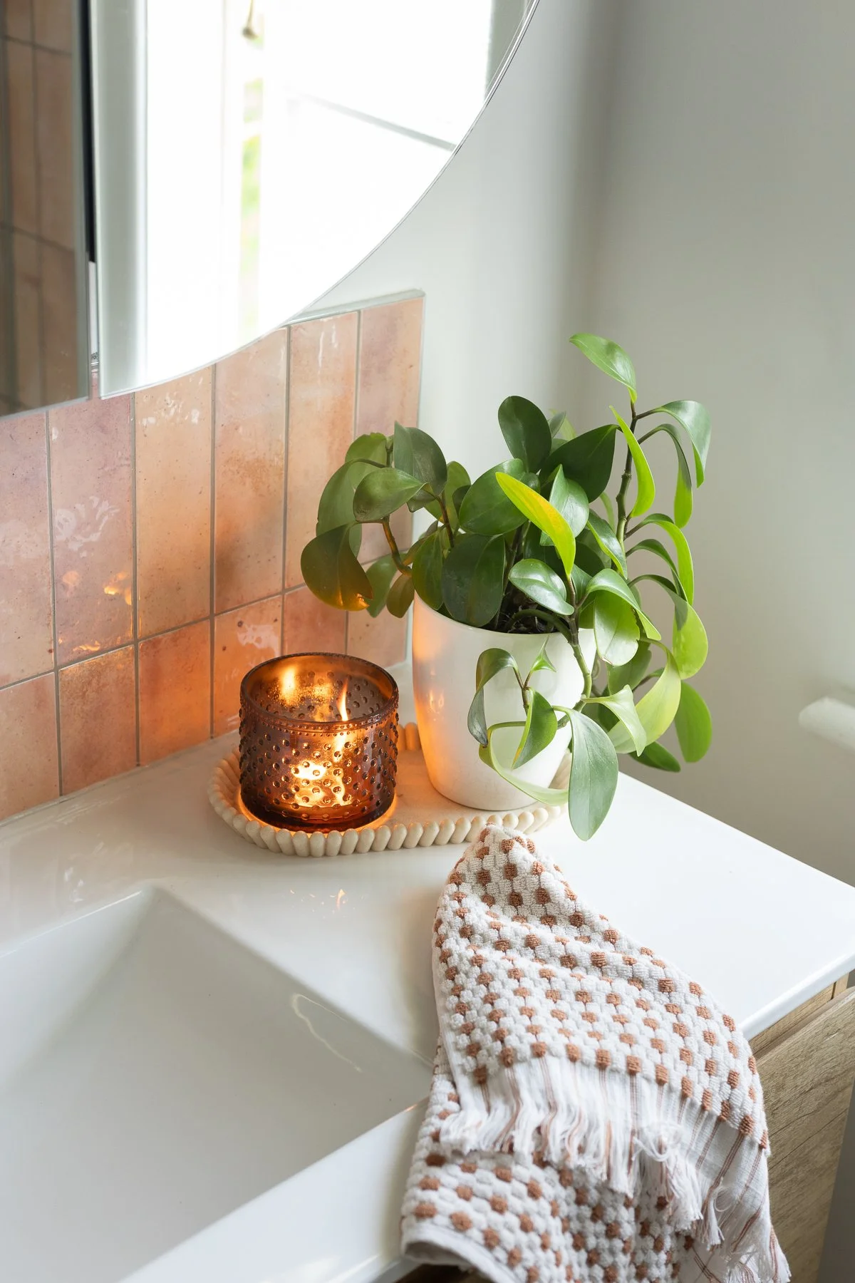 A bathroom sink area with a potted plant, a lit candle in a brown perforated candle holder, a white ceramic tray, and a patterned towel draped over the edge of the sink.