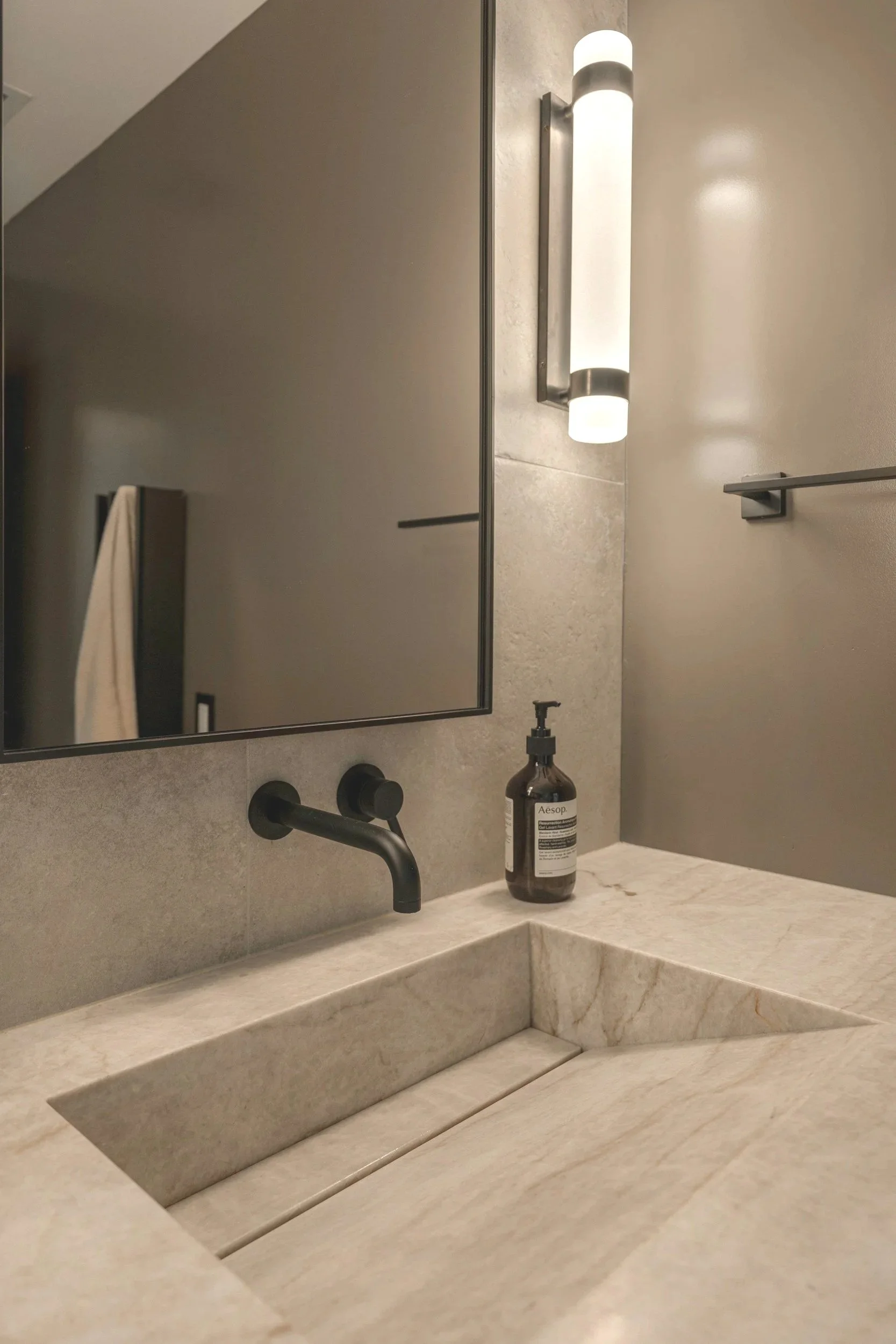 Bathroom countertop with a marble surface, a square undermount sink, a black faucet, a mirror, a wall-mounted light fixture, and a bottle of Aesop hand soap.