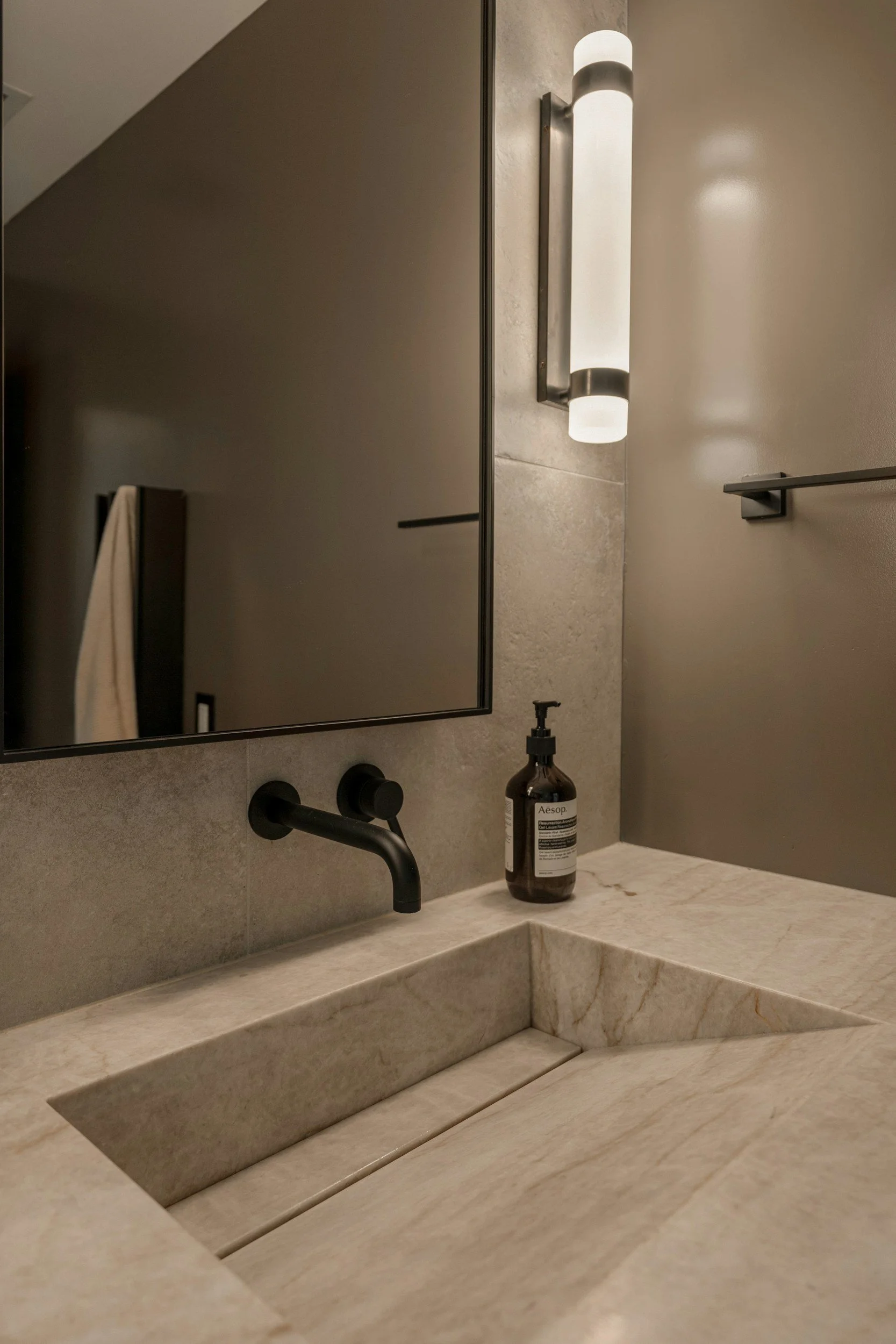 Close-up of a bathroom backsplash and sink with a bottle of Aesop hand soap, black faucet, mirror, wall light, and beige countertop.