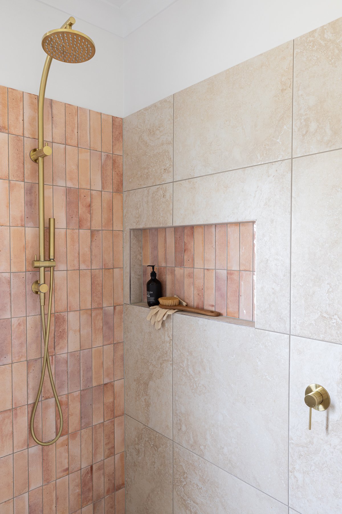 Shower with gold fixtures, pink and beige tiles, a built-in niche with a black soap dispenser, a wooden brush, and a pair of gloves.