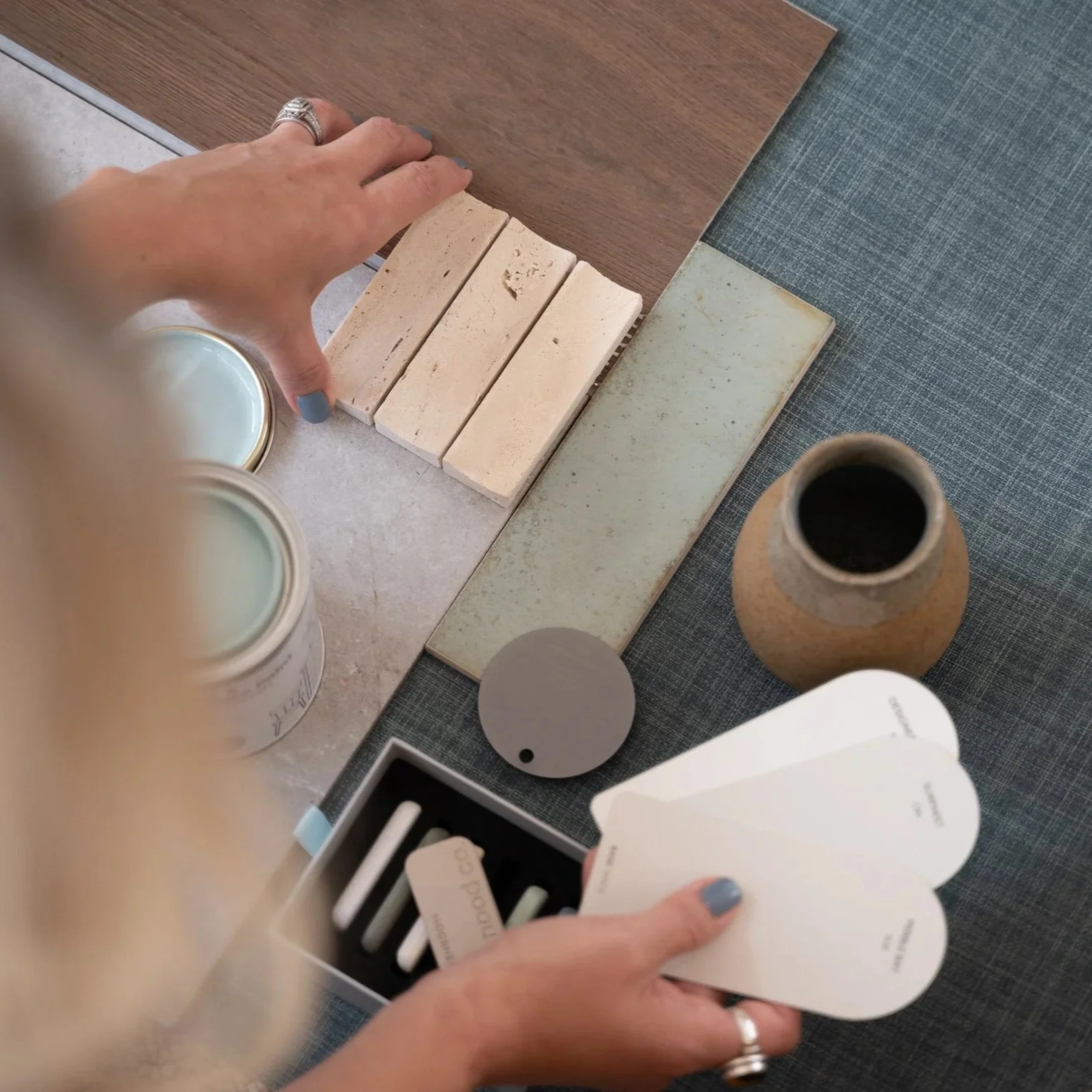 A person's hand with painted blue nails holding a fan-shaped color palette with various shades and names of paint colors. On the table, there are paint cans, tiles, and a small round mirror or compact.