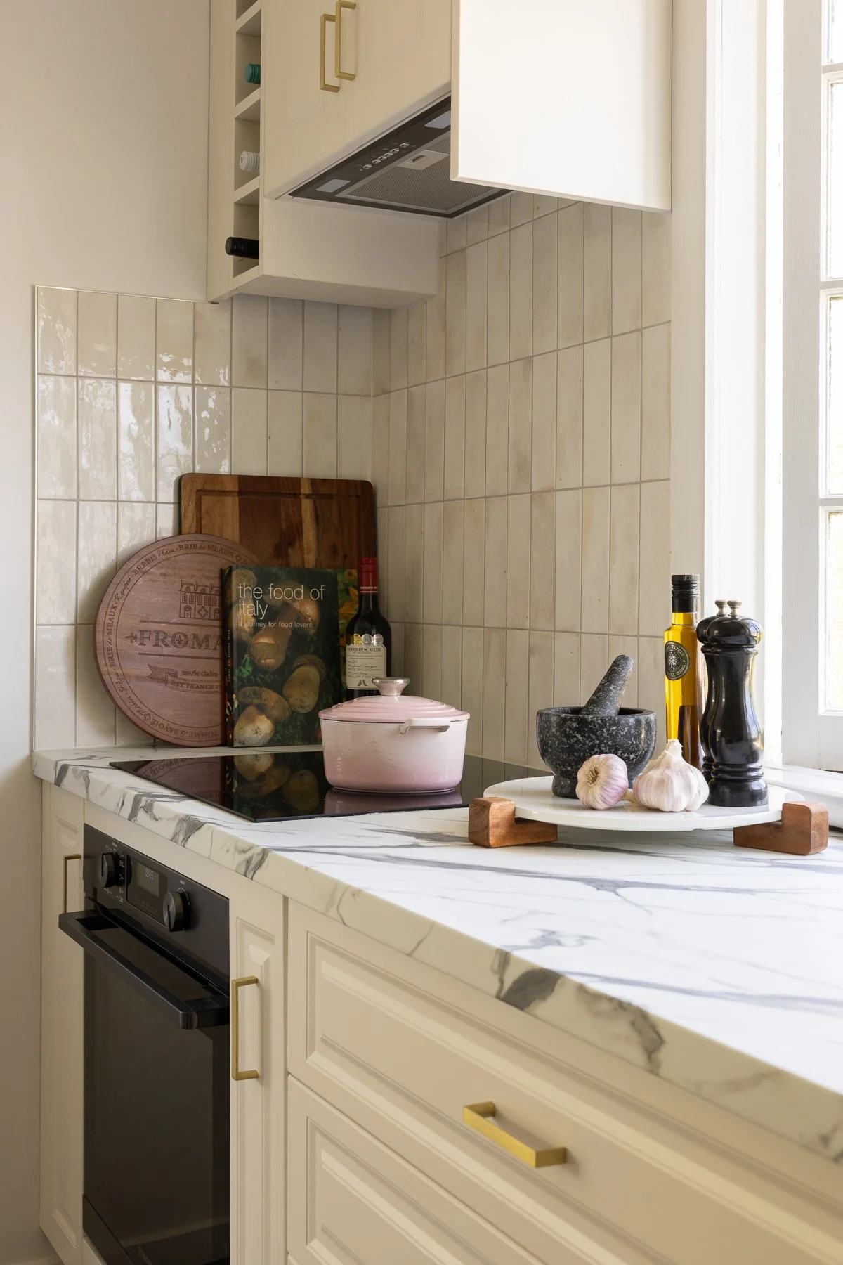 A modern kitchen with white cabinets and marble countertop. On the counter, there is a round black stone mortar and pestle, garlic, olive oil, salt and pepper shakers, a pink casserole dish, a wooden cutting board, and two recipe books titled "the fo