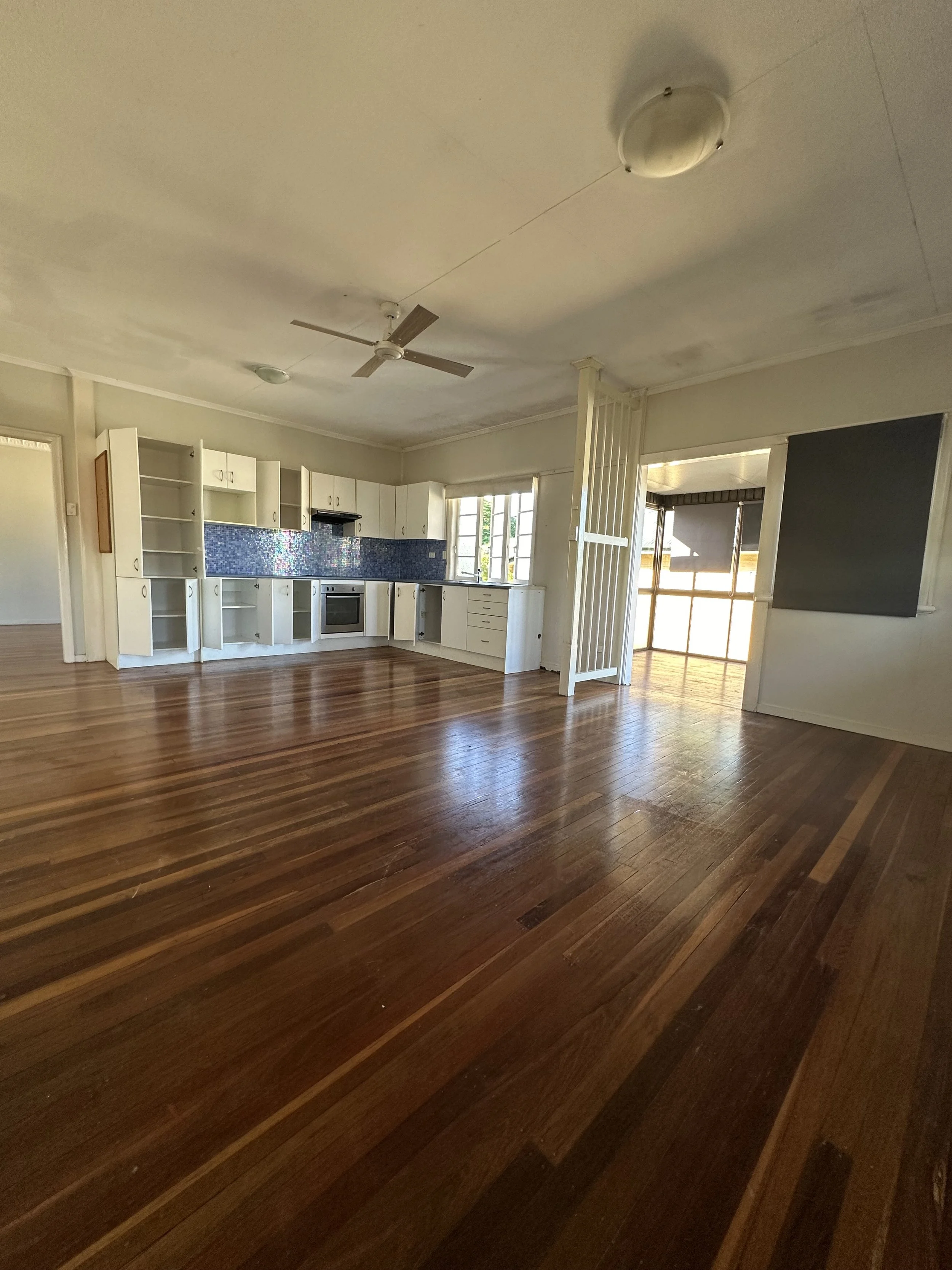 Empty unrenovated living room with hardwood floors, old white kitchen cabinets, and a ceiling fan.