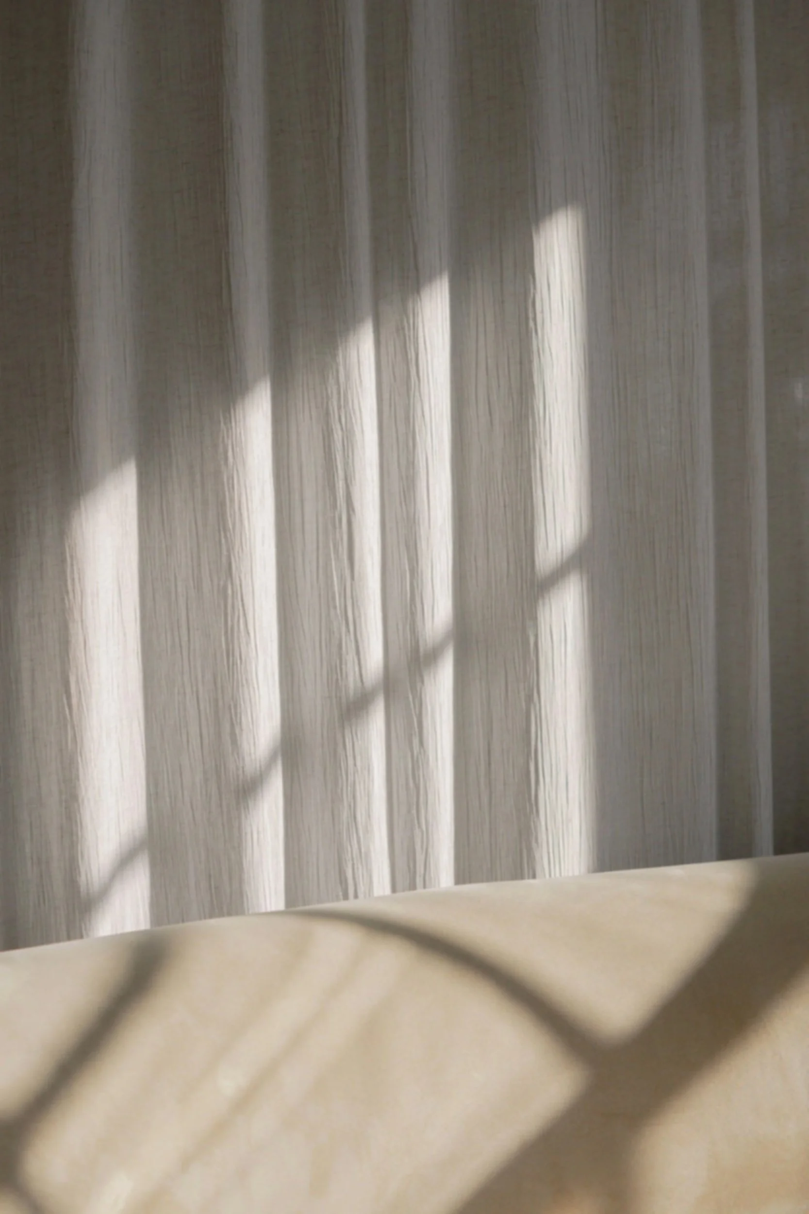 Sunlight creates shadows through window blinds onto a beige surface and a light-colored curtain.