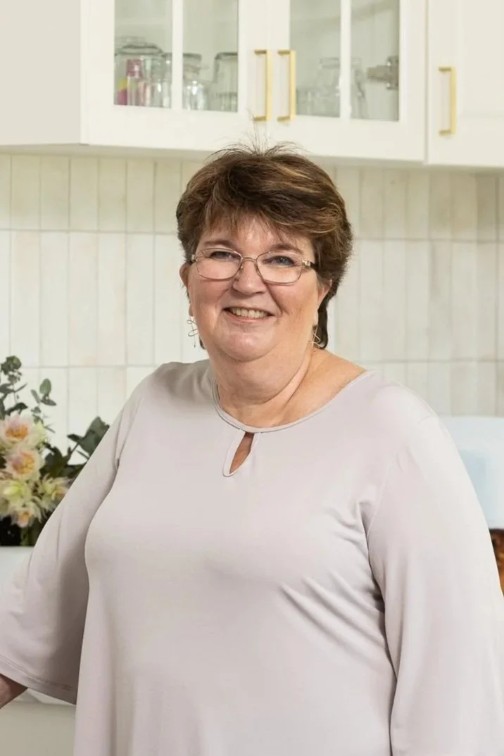 Director, Sue smiling and standing in a kitchen with white cabinets and a floral arrangement in the background.