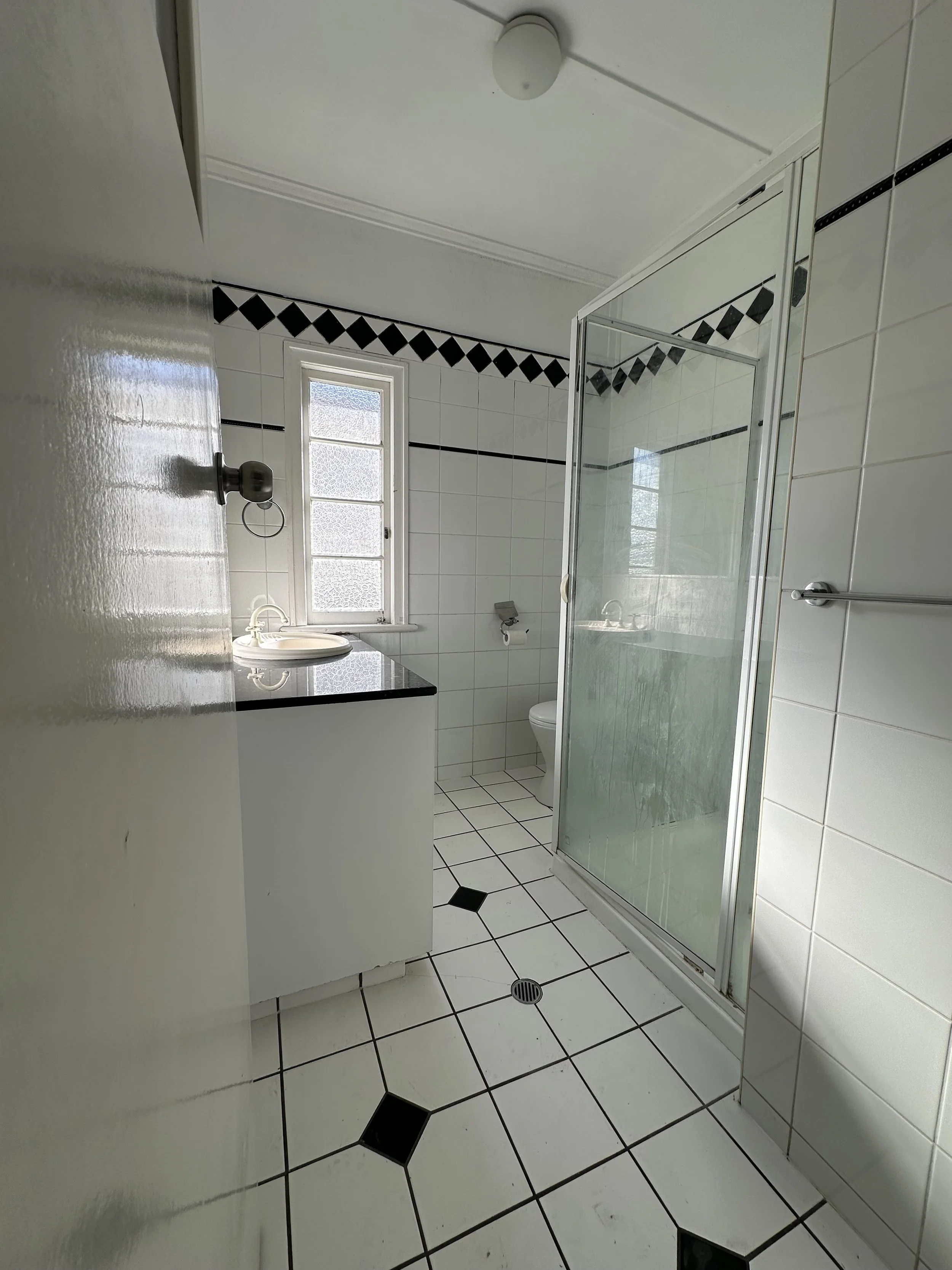 Unrenovated bathroom with dated white tiles, a glass shower enclosure, a small window with frosted glass, a white toilet, and a white sink with a black countertop.