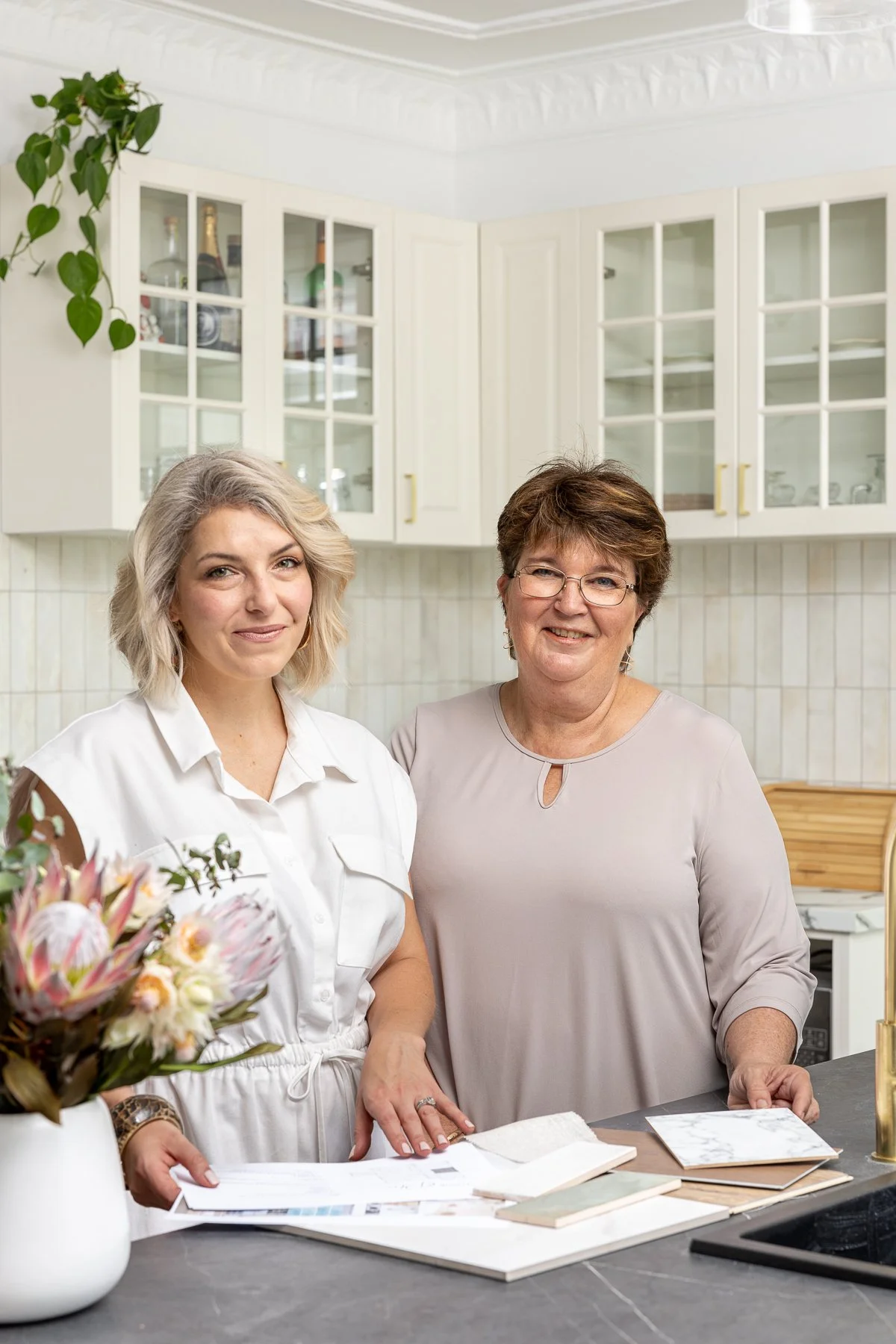 Directors Lisa and Sue standing at a marble countertop in a bright, white kitchen with glass-front cabinets, holding papers and smiling.