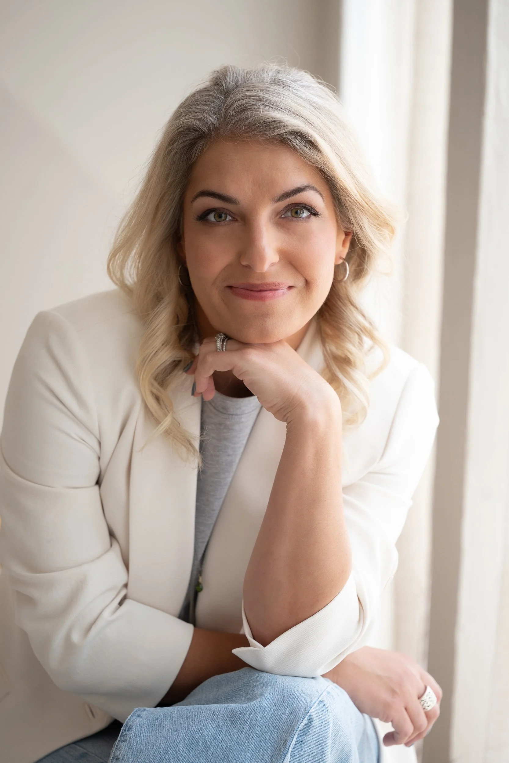Director, Lisa smiling gently, resting her chin on her hand, wearing a white blazer and light-colored top, sitting indoors near a window with natural light.