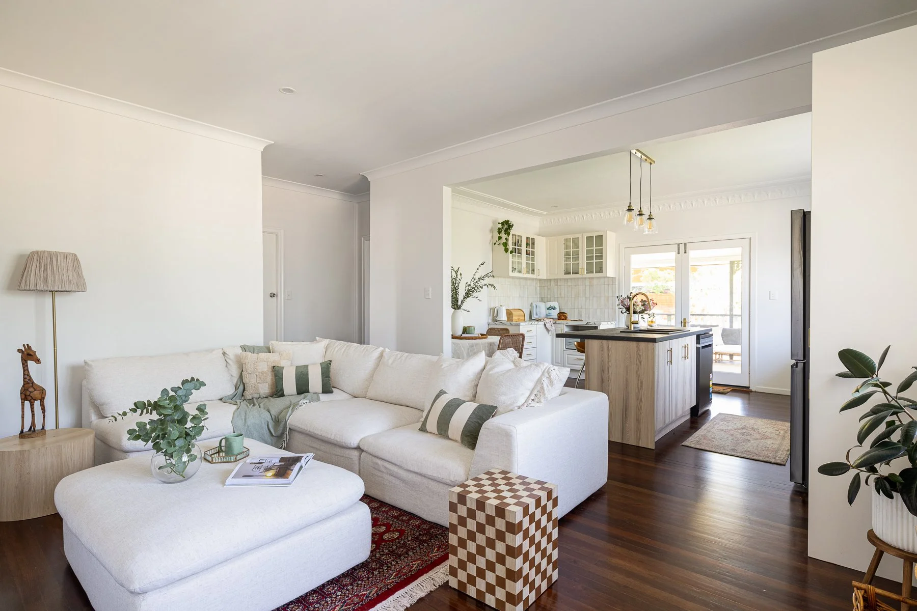 Modern living room with a white sectional sofa and green striped pillows, next to a wooden side table with a giraffe decor and a floor lamp, overlooking an open kitchen with white cabinets and a wooden island, hardwood floors, and sliding glass doors