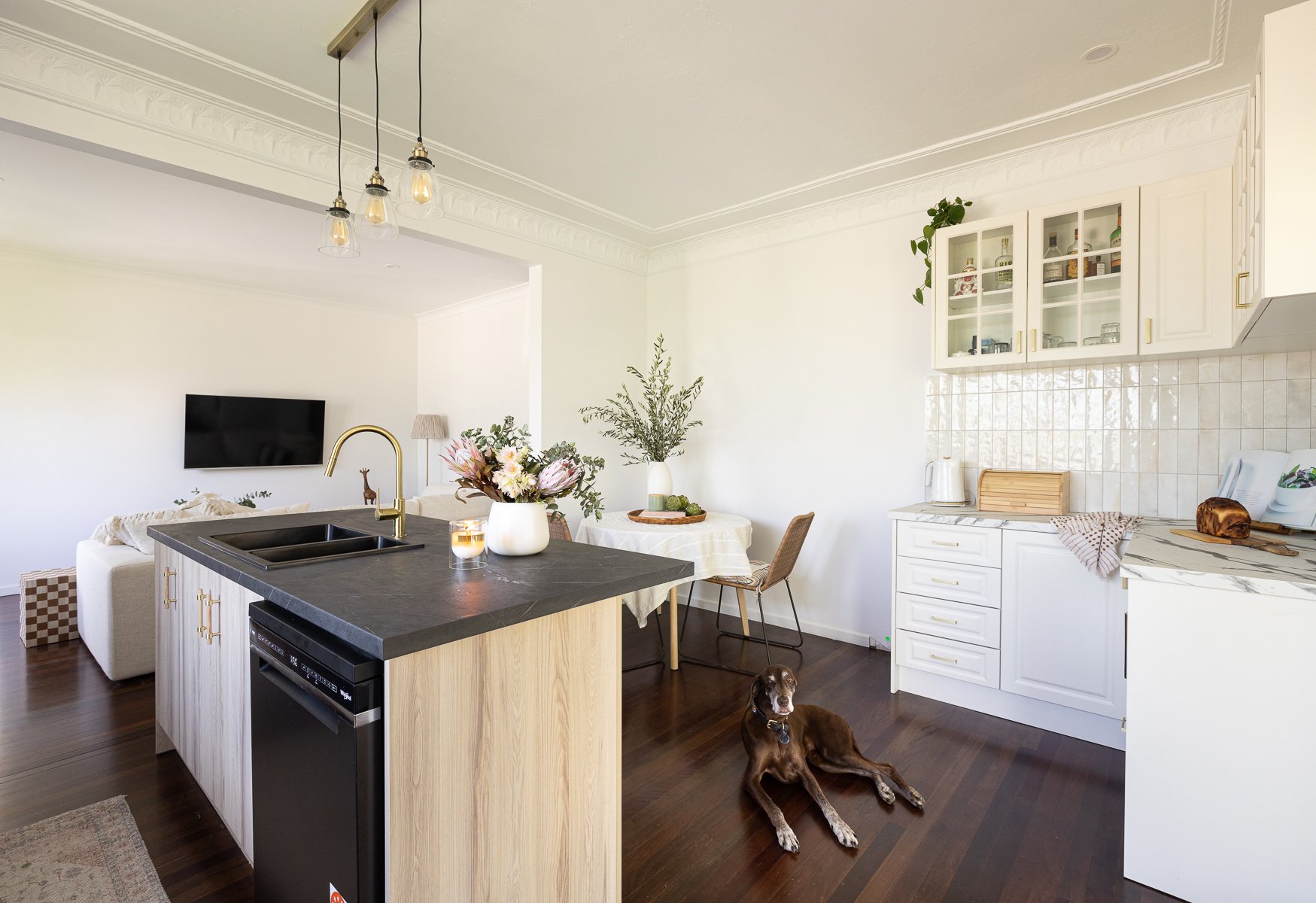 Open-concept kitchen and living room with white cabinets, a black countertop island, a dining table with a floral centerpiece, a brown dog lying on the dark wood floor, and decorative plants.