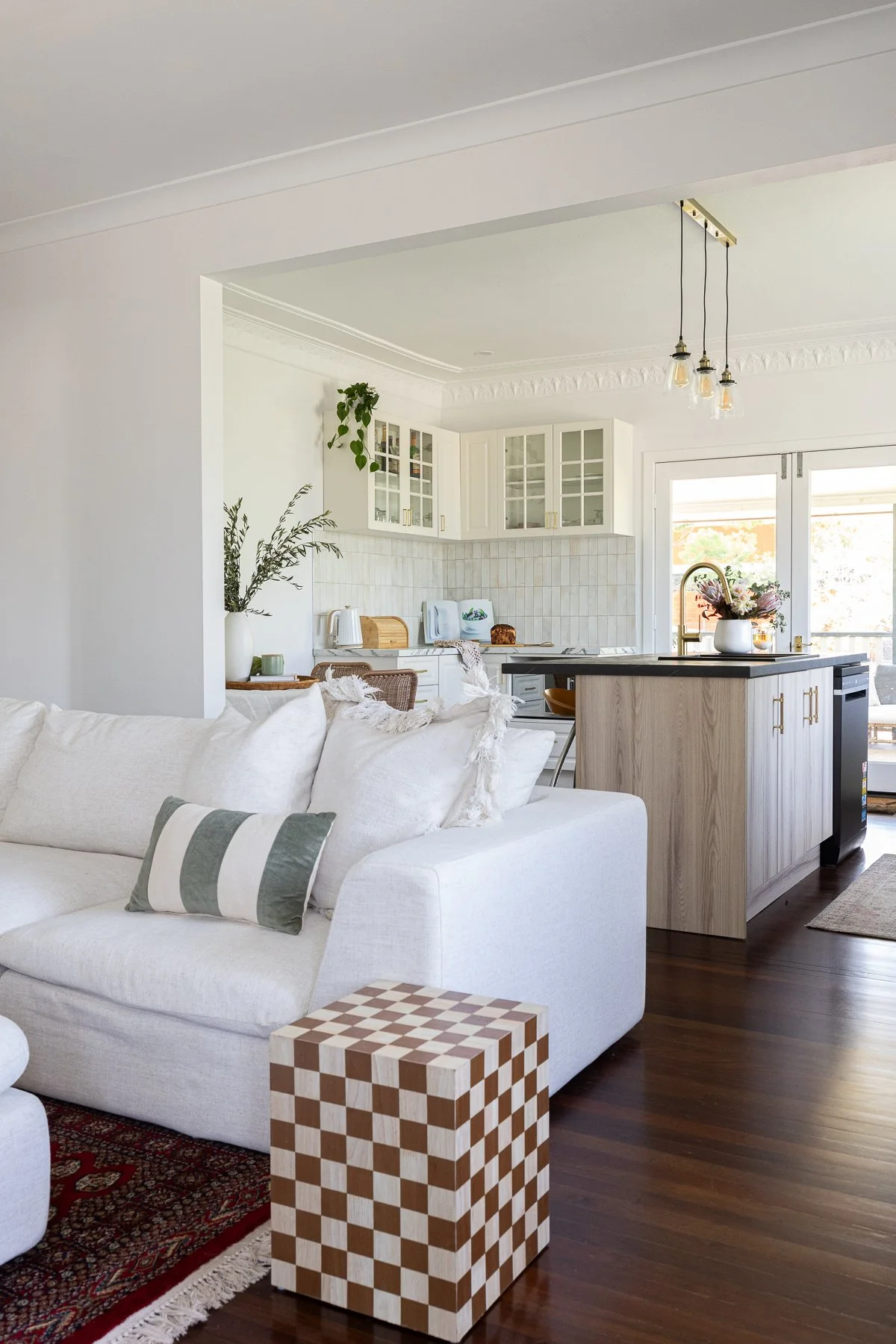 A bright, modern living room and kitchen with a white sofa, patterned pillow, wooden cube table, and a view into the kitchen with white cabinets, hanging light fixtures, plants, and a sliding glass door.
