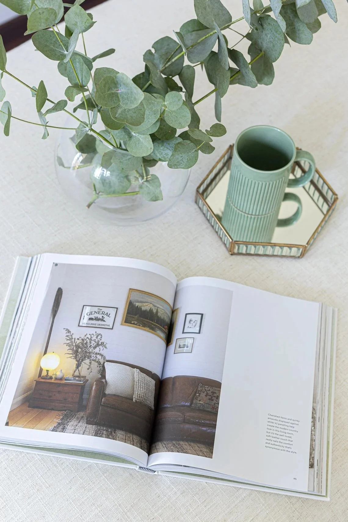 A tabletop with a vase of eucalyptus leaves, a green mug on a decorative tray, and an open magazine showing a living room interior.