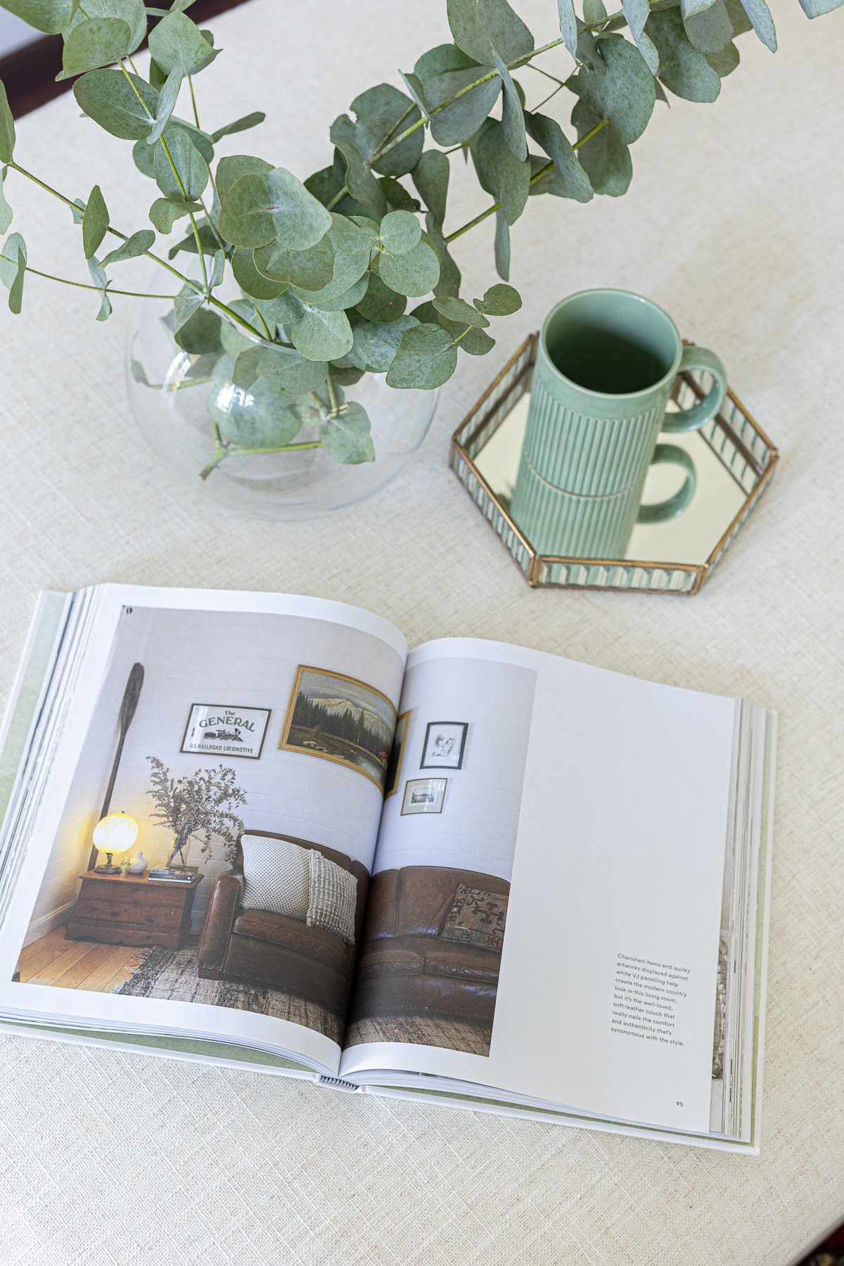 Close-up of an open magazine displaying a living room interior with a brown leather sofa, framed artwork on the wall, and a wooden side table with a lamp and a vase of flowers. The magazine is on a white textured surface.