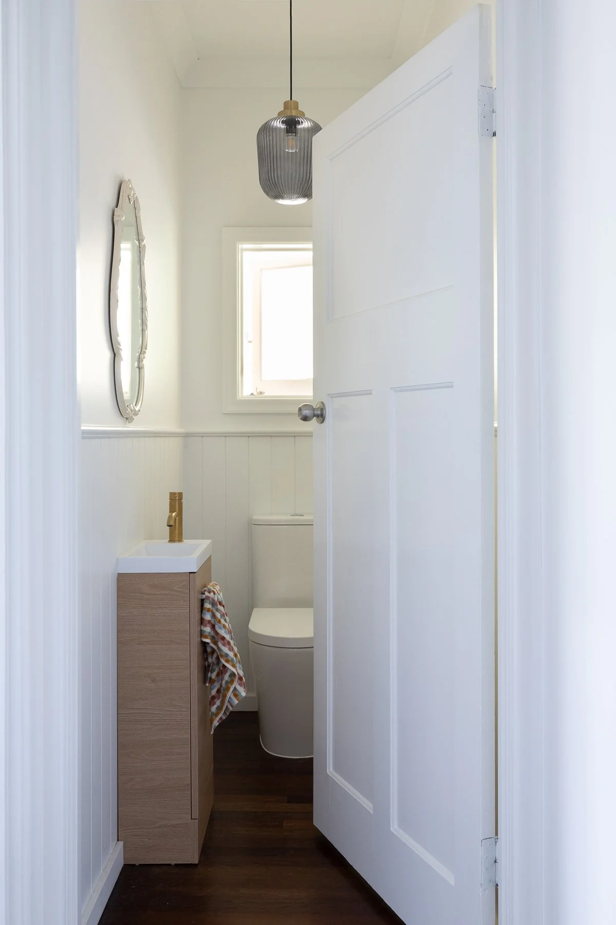 A small, modern, minimalist half-bathroom with white walls, a white door partially open, a small sink with a wood cabinet, a towel hanging on the side, a ceiling light fixture, a window with natural light, a toilet, and a decorative mirror on the wal
